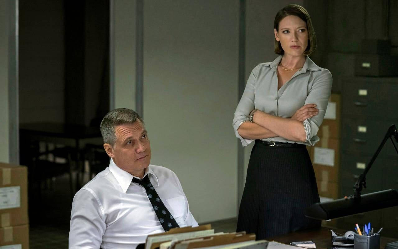 Netflix《Mindhunter》溫迪·卡爾與比爾·坦奇 壁紙 — 一個緊張的辦公室場景，一名身穿白襯衫打領帶的男子坐在辦公桌前，而一名身穿灰色襯衫的女子雙手交叉站立，嚴厲地看著他。