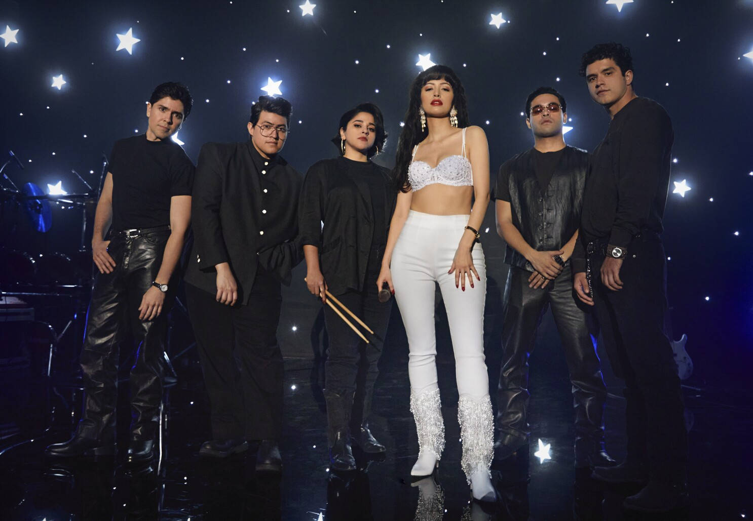 Netflix《Selena: The Series》劇組群像 壁紙 — 一位穿著白色亮片褲和胸罩上衣的迷人女歌手在星光燈光下站在舞台中央，被她的樂隊包圍。