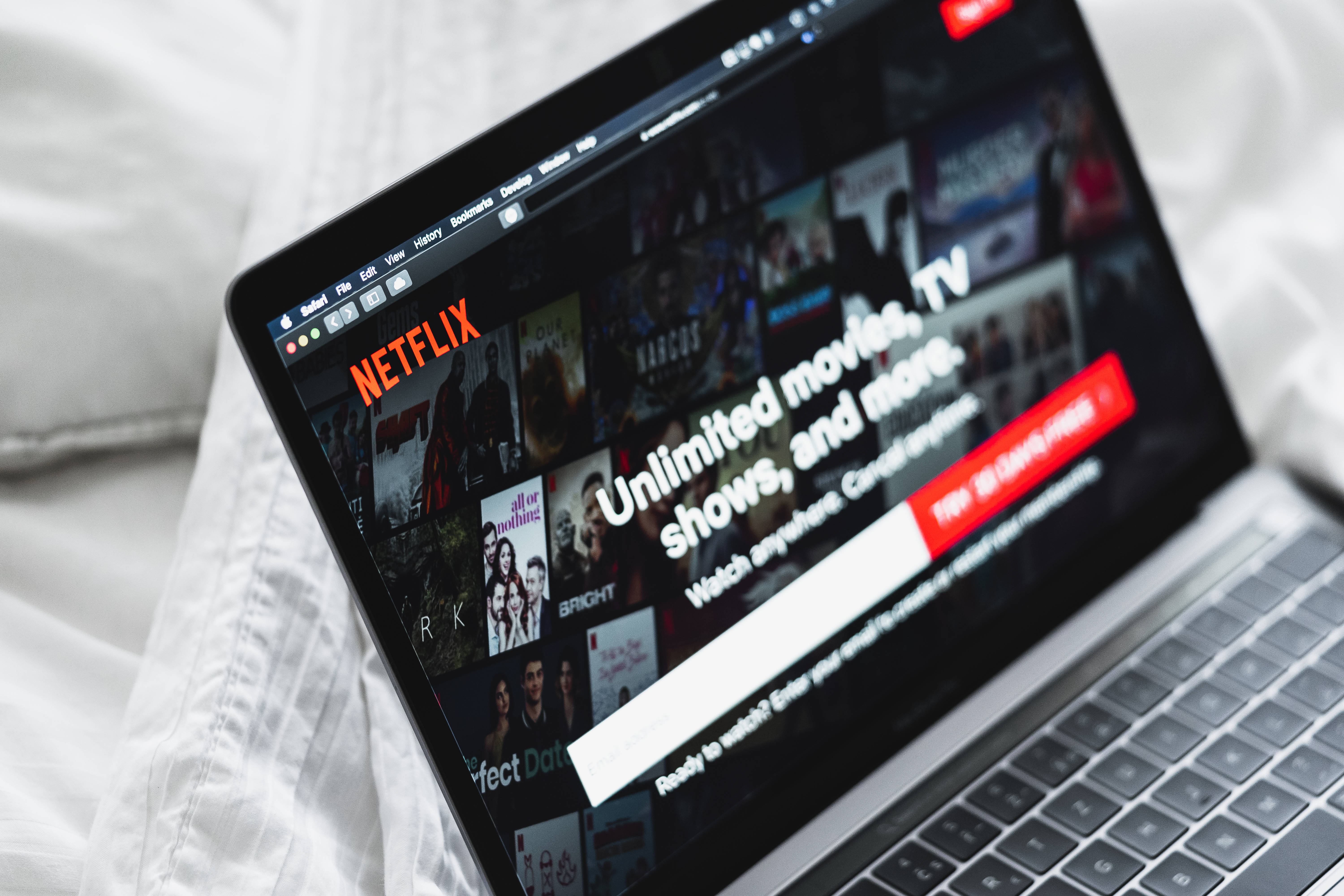 筆記本電腦螢幕顯示Netflix網站 壁紙 — 一部顯示Netflix主頁的筆記型電腦放在柔軟的白色床單上，營造出舒適的室內氛圍。