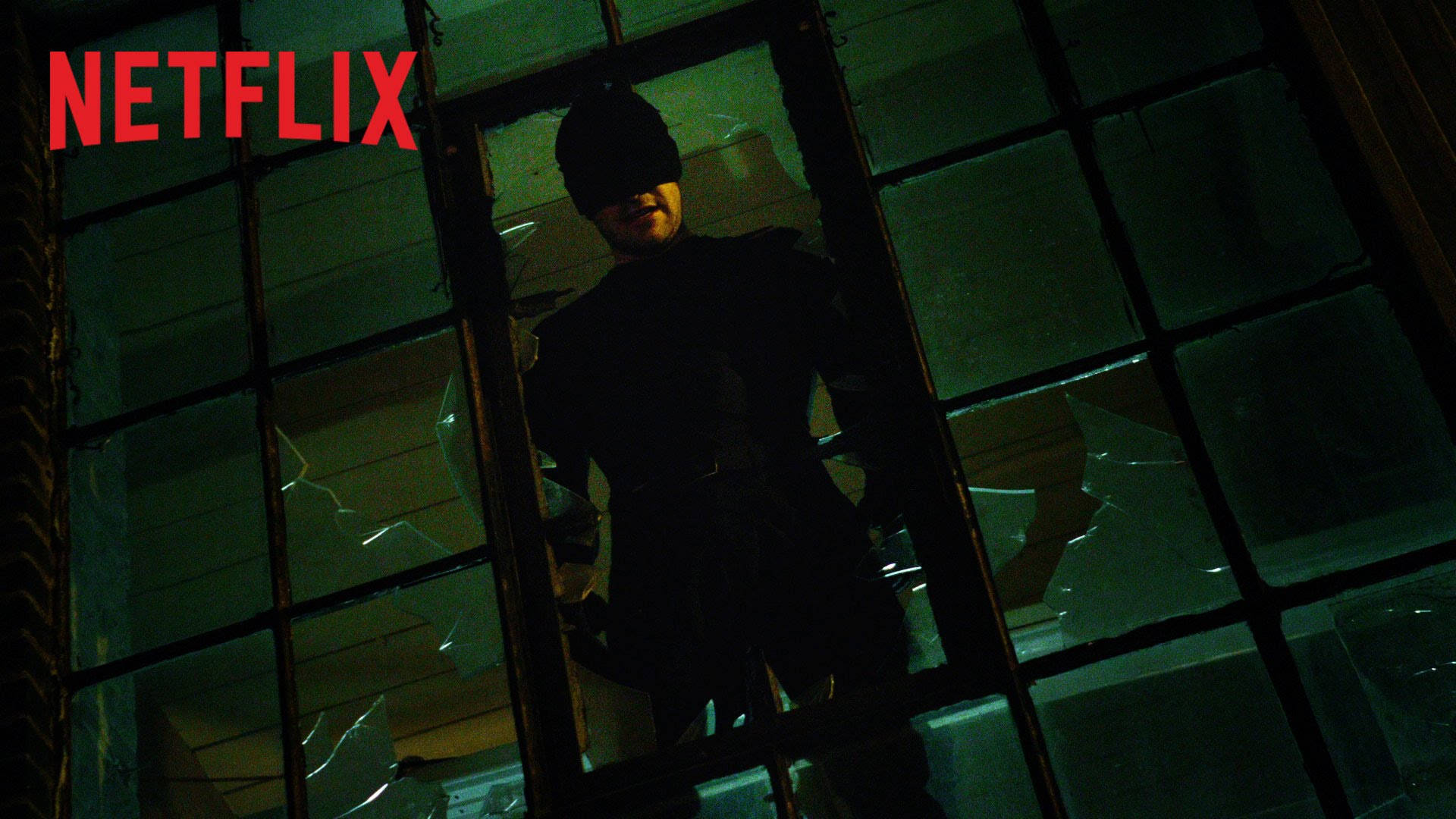 Netflix《夜魔俠》窗邊場景 壁紙 — 一名戴面具的義警站在破碎的窗戶後面，處於昏暗的城市環境中，帶有綠色調的燈光。