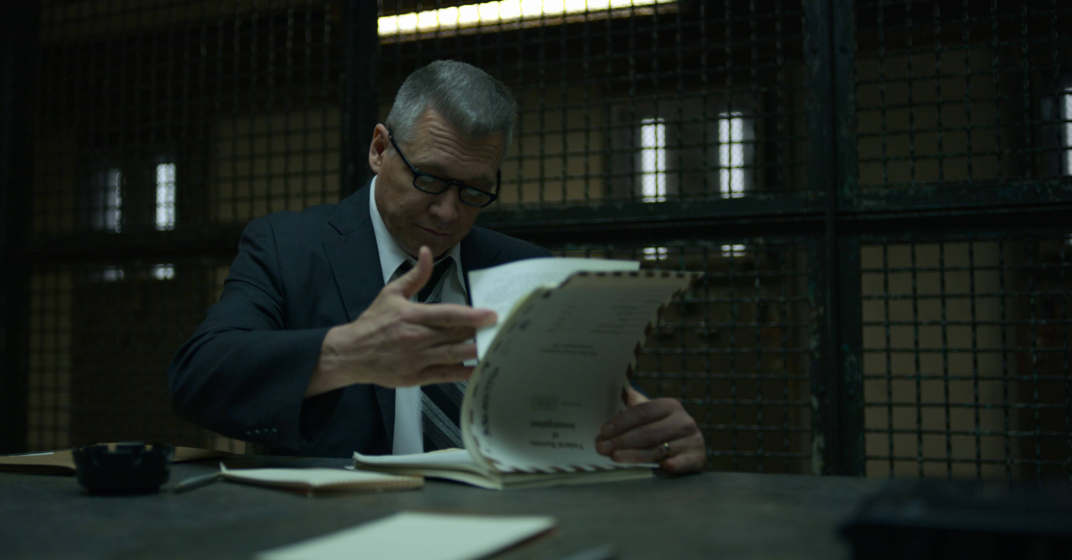 Netflix《心理追兇》盤問場景 壁紙 — 一名身穿西裝、戴着眼鏡的男子在昏暗的審訊室裏，隔着鐵欄杆在桌上專注地審閱文件。