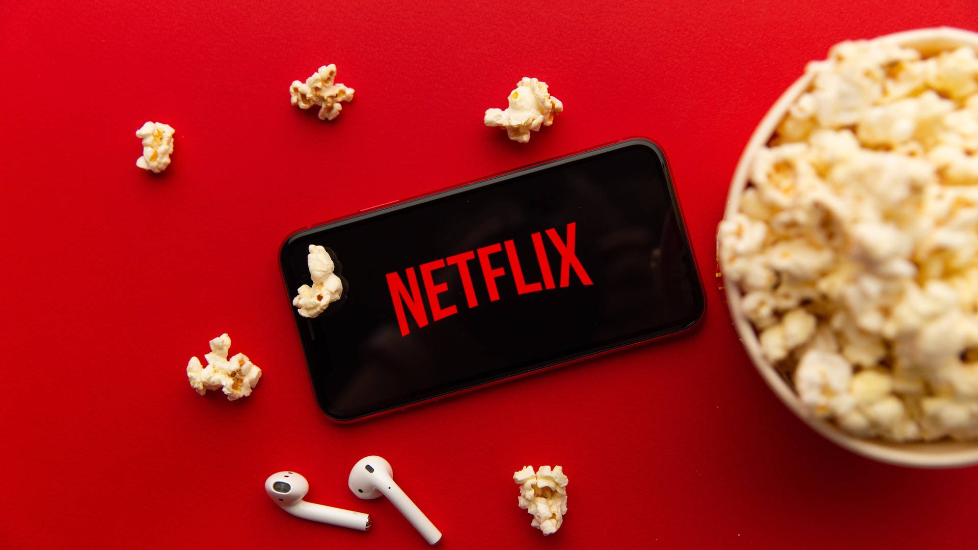 Netflix與爆米花 壁紙 — 一部顯示Netflix標誌的智能手機放在鮮紅色的表面上，周圍散落著爆谷和白色無線耳機。