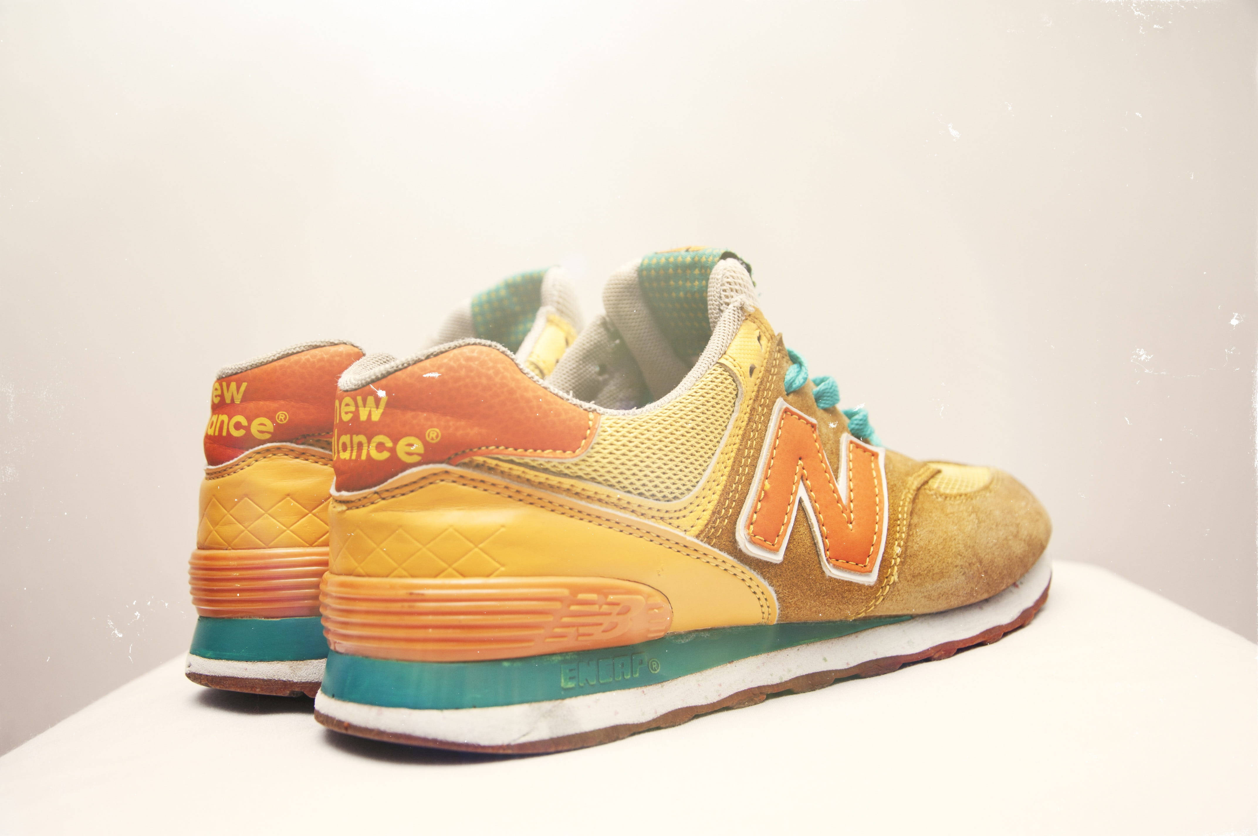 New Balance 後側視角 壁紙 — 一對復古New Balance運動鞋，採用溫暖的橙黃色和藍綠色調，在柔和的白色背景上展示。