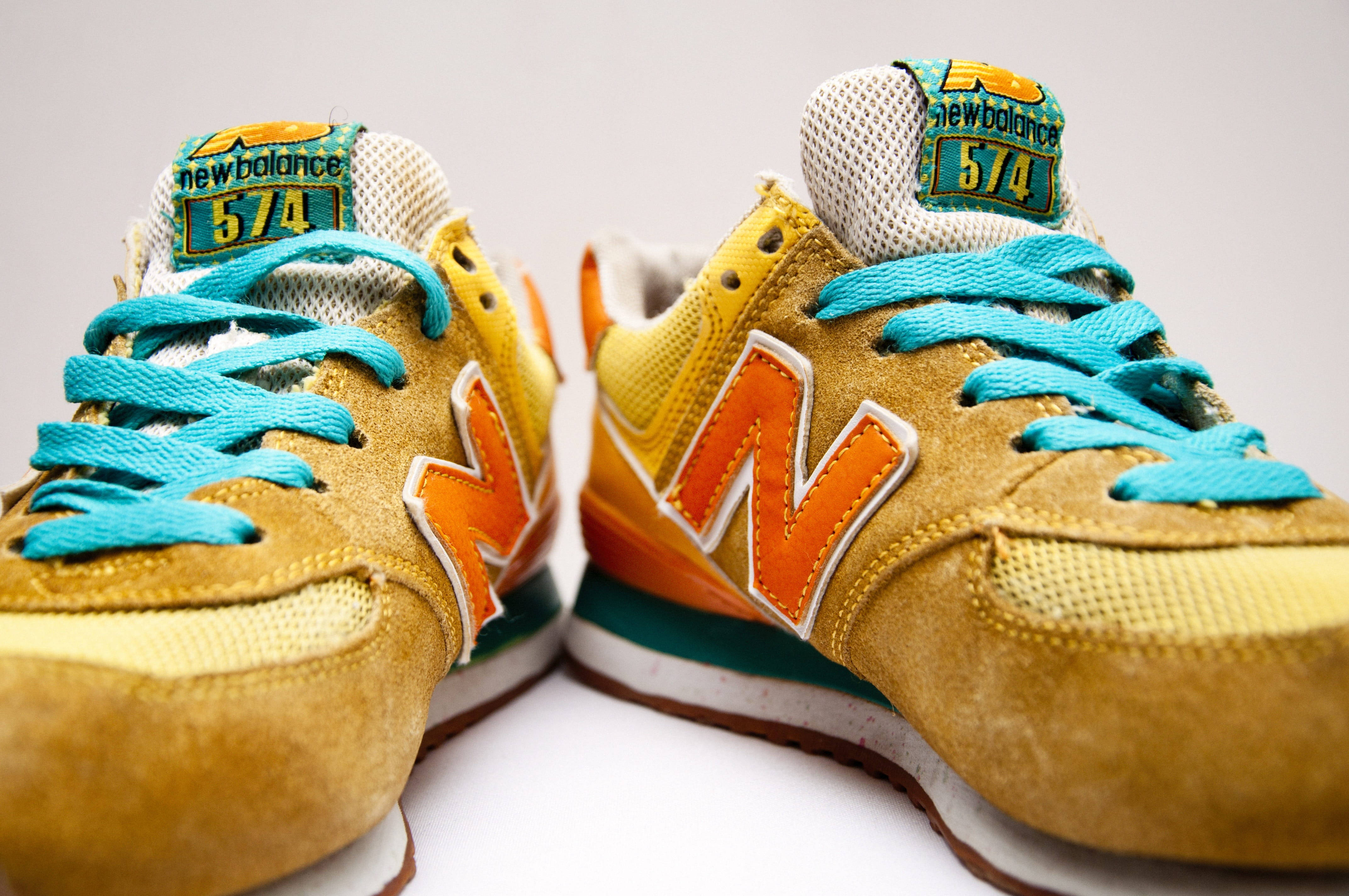 New Balance 橘黃鞋款 壁紙 — 鮮黃色和橙色New Balance 574運動鞋的特寫，配有藍綠色鞋帶，背景為純白色。