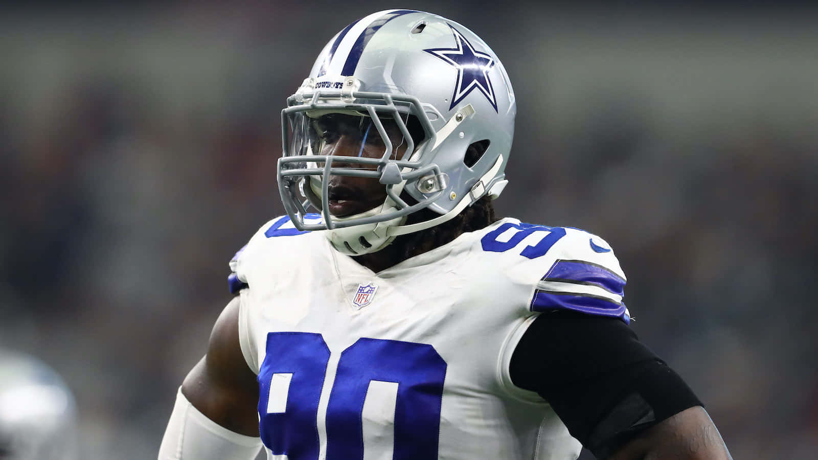 NFL運動員德馬庫斯·勞倫斯 壁紙 — 達拉斯牛仔隊美式足球員身穿白色和藍色球衣，背號90，在球場燈光下比賽時凝視前方。