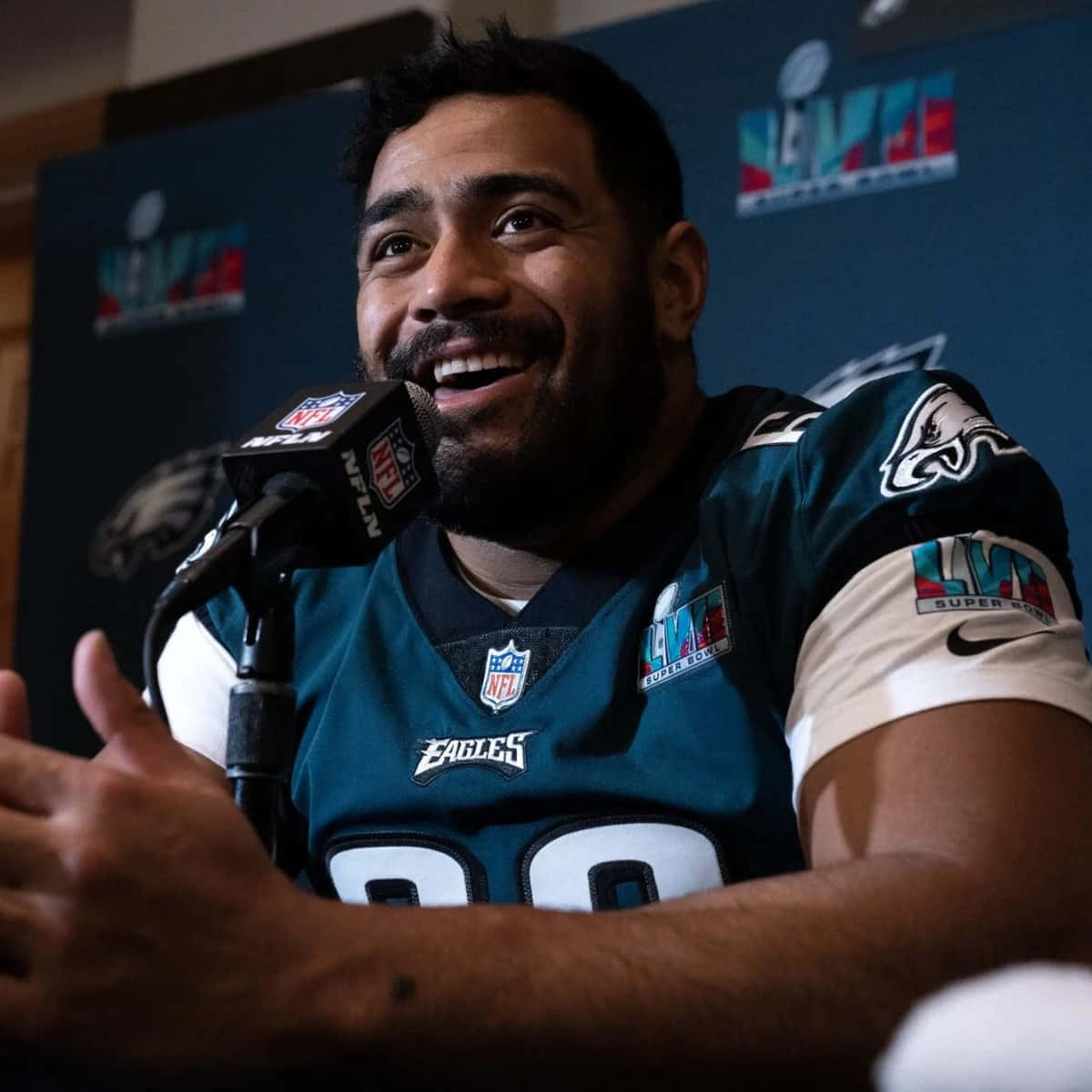  壁紙 — 一位面帶微笑的NFL美式足球員身穿費城老鷹隊球衣，在記者會上對著麥克風發言。