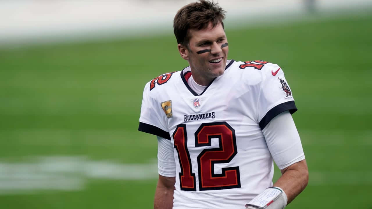 追蹤最新NFL與足球新聞 壁紙 — 一位微笑的坦帕灣海盜隊四分衛，身穿白色12號球衣，站在綠色足球場上。