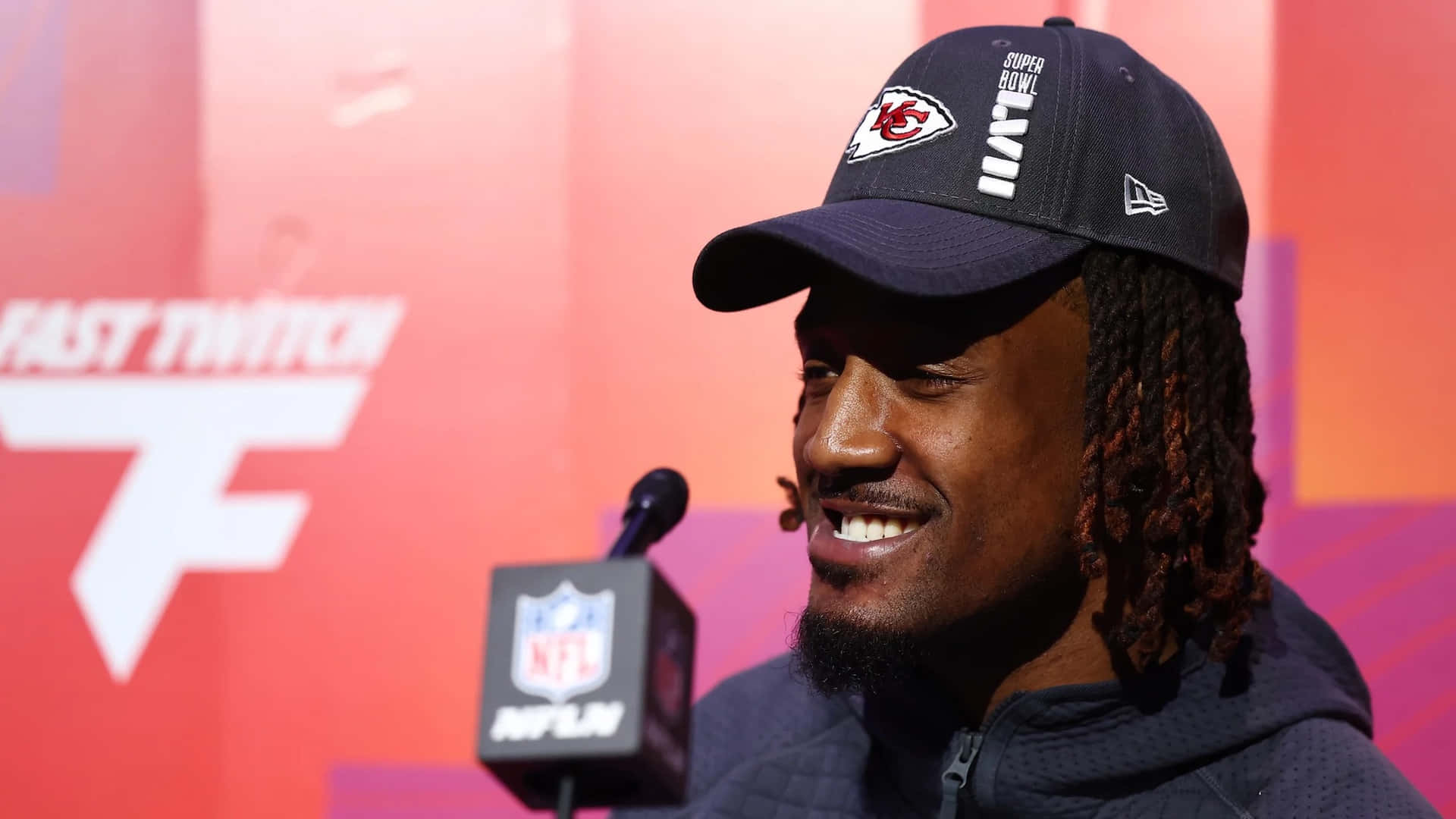  壁紙 — 一位面帶微笑的NFL球員戴著酋長隊帽子，在Twitch活動中發言，現場有品牌麥克風和鮮紅背景。
