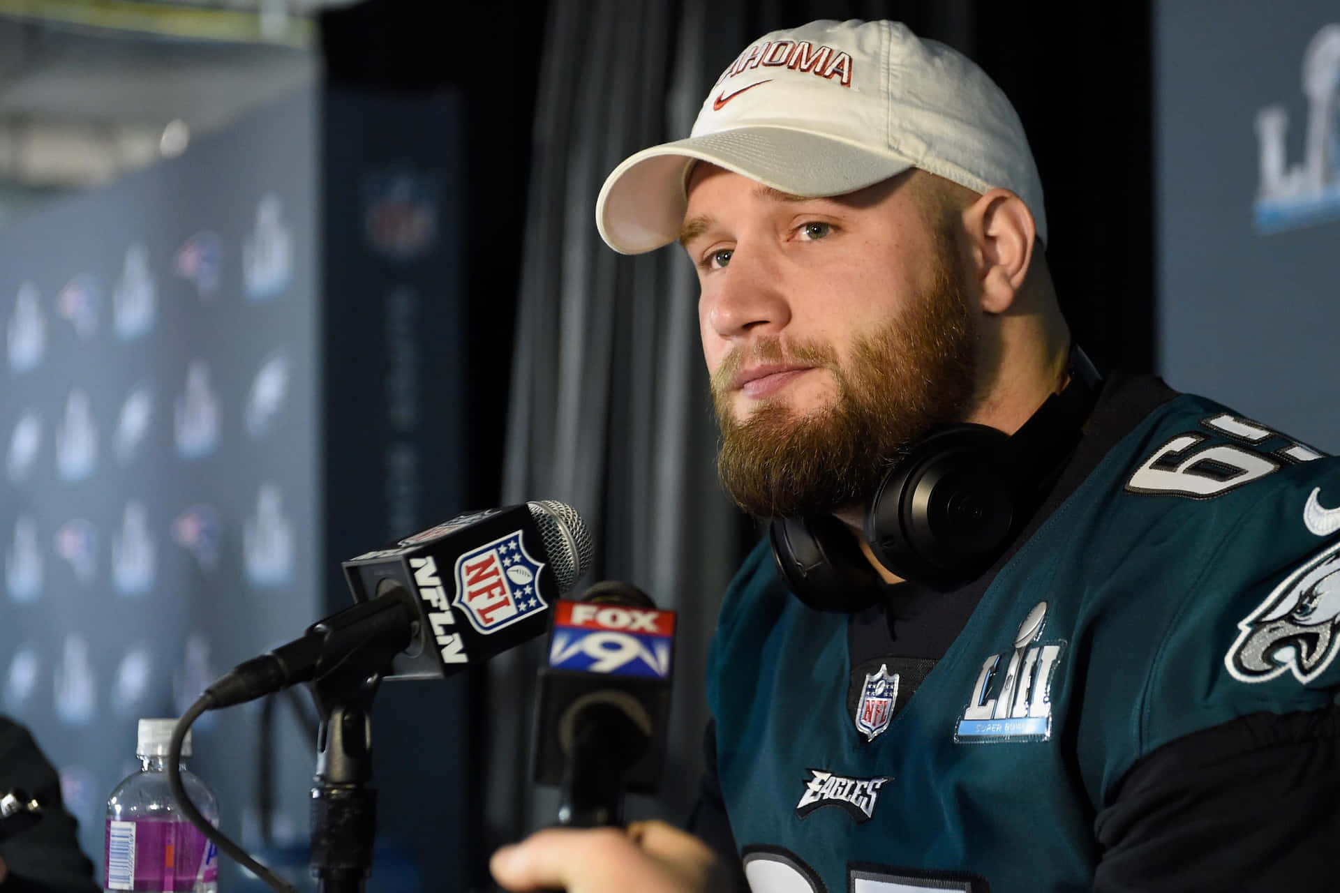  壁紙 — 一位留鬍鬚的NFL球員，身穿費城老鷹隊球衣和帽子，坐在記者會上，面前有麥克風。