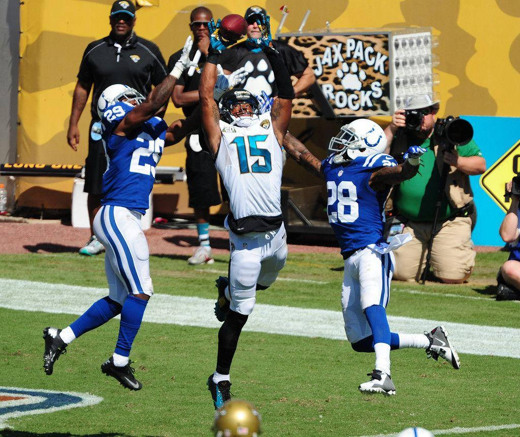 NFL 阿倫·羅賓遜 杰克遜維爾美洲虎 與底特律獅隊比賽 壁紙 — 一名身穿白色15號球衣的美式足球員跳起接球，兩名藍色球衣的防守球員在陽光明媚的球場上追趕他。
