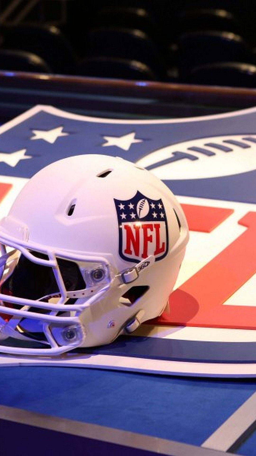 NFL 標誌印於足球頭盔 壁紙 — 一個純白色的NFL美式足球頭盔放在色彩繽紛的球隊標誌地板上，背景是模糊的體育館座位。