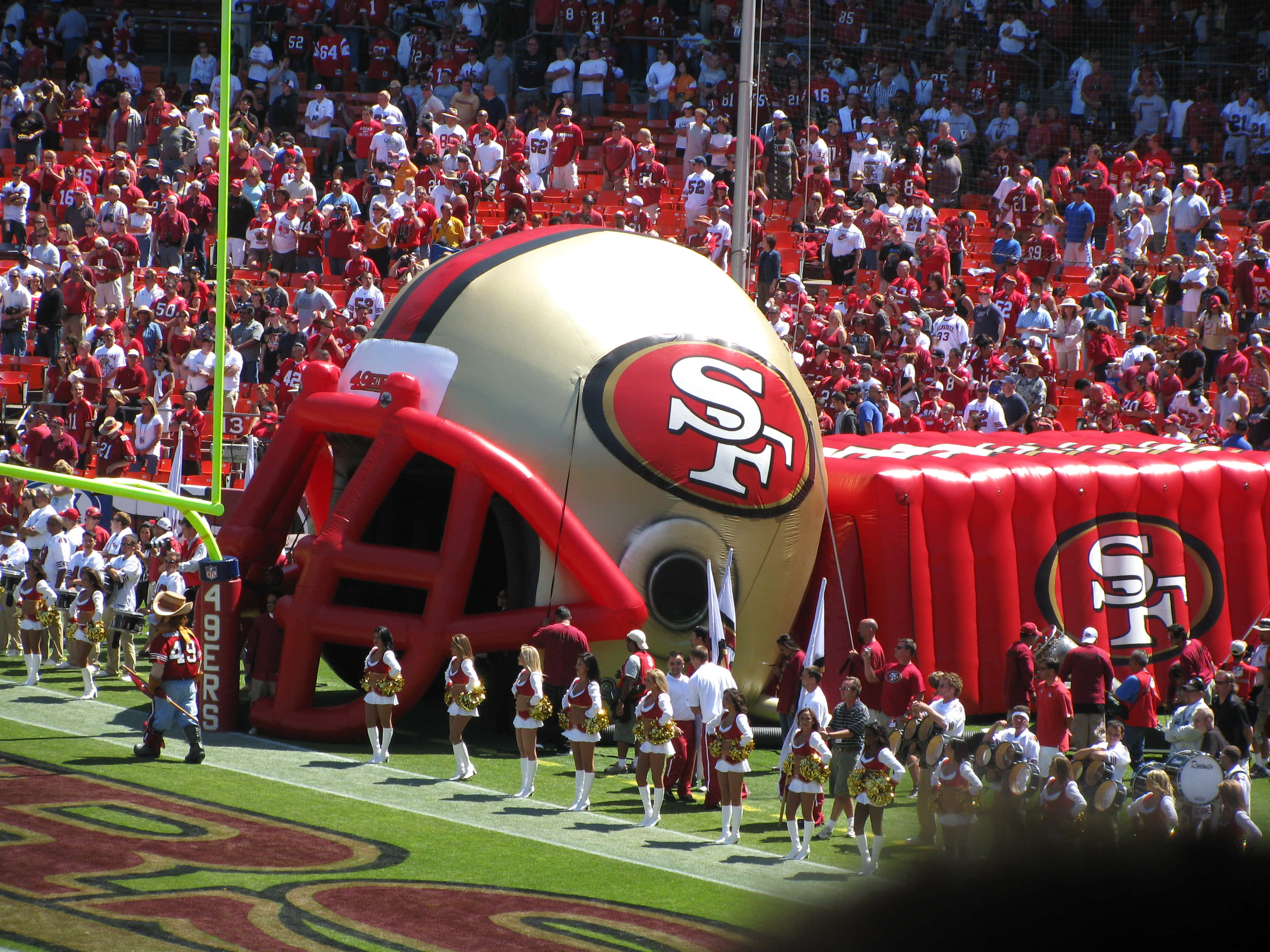NFL傳奇球星集錦 壁紙 — 一個巨大的三藩市四九人隊充氣美式足球頭盔在陽光明媚的球場上佔據主導地位，啦啦隊在比賽期間列隊走過。