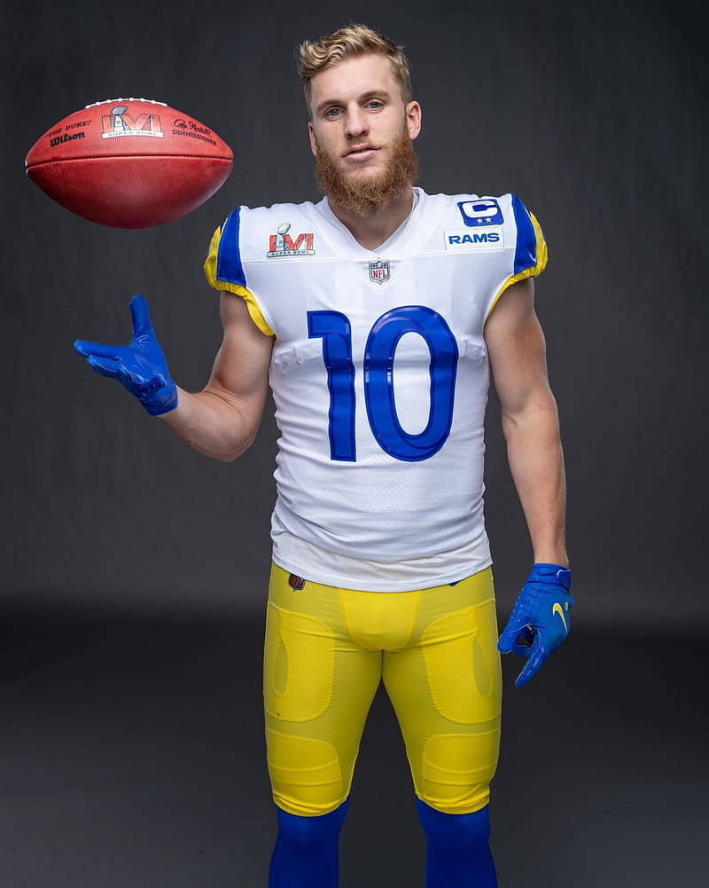NFL洛杉磯公羊隊庫珀·庫普攝影棚拍攝 壁紙 — 一位留鬍鬚的美式足球員，身穿白色、藍色和黃色的公羊隊球衣，在深色攝影棚背景前，用戴手套的手平衡一個紅色美式足球。