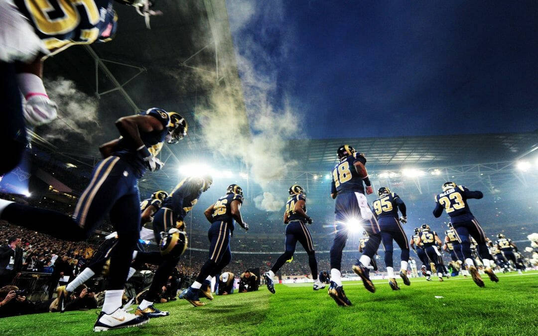 NFL球員在球場奔跑 壁紙 — 身穿藍色和金色球衣的足球運動員在體育場燈光下衝入球場，身後煙霧翻騰。
