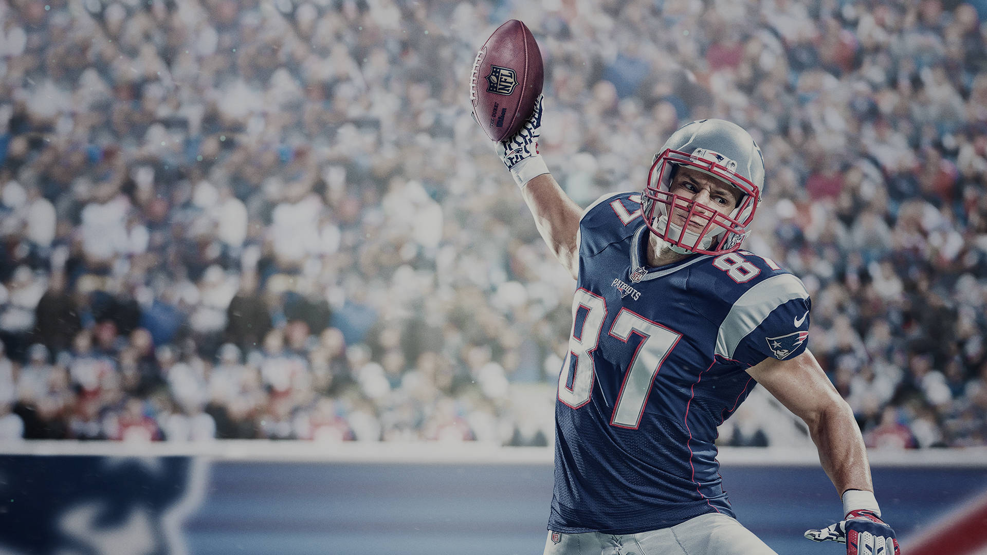 NFL球員投球瞬間 壁紙 — 一名身穿藍色球衣的美式足球員，在模糊的體育場人群前，勝利地將一個棕色橄欖球高舉過頭頂。