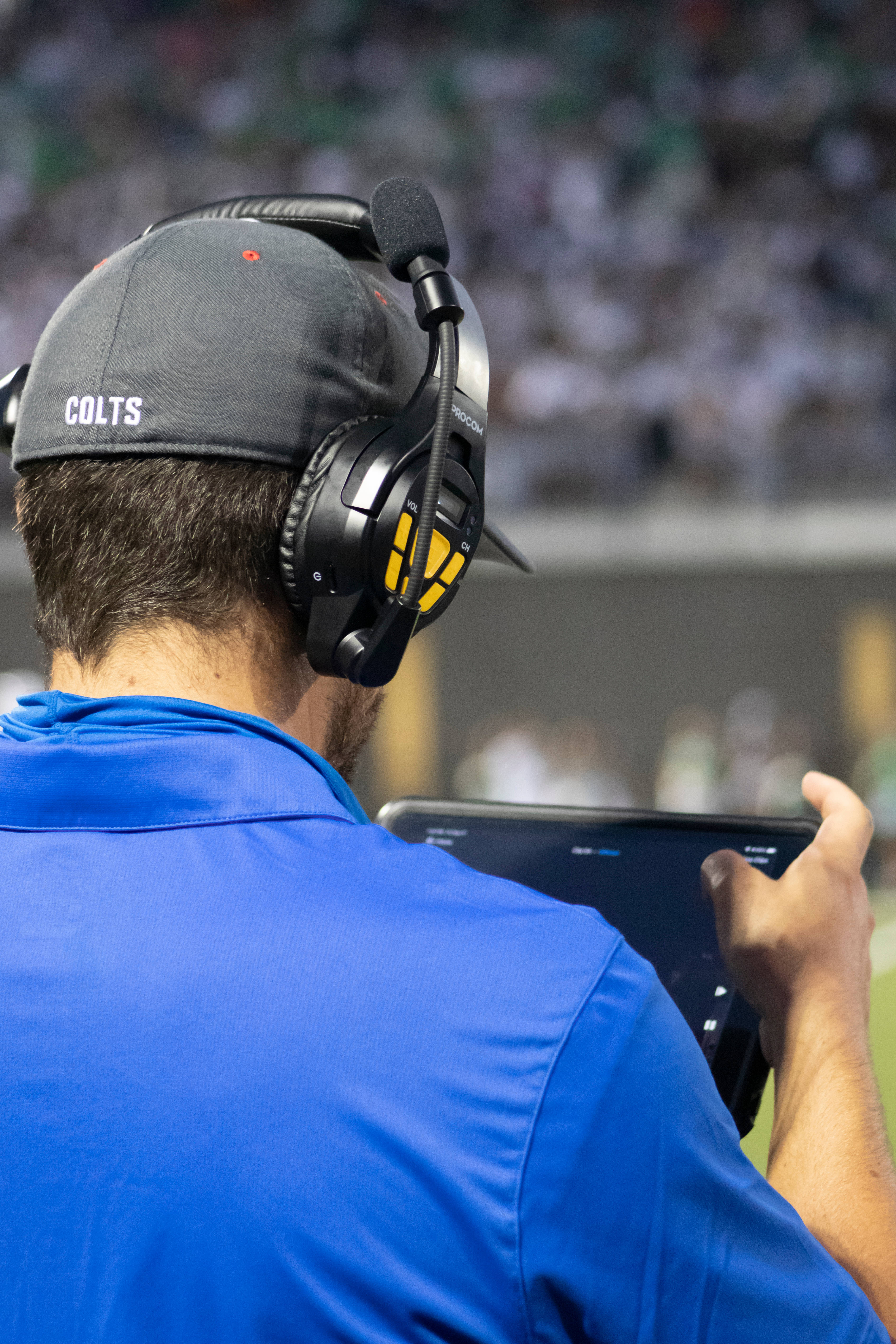 NFL美式足球教練 壁紙 — 一位戴著Colts帽和耳機的教練在足球比賽中專注地看著平板電腦，背景是模糊的體育場人群。