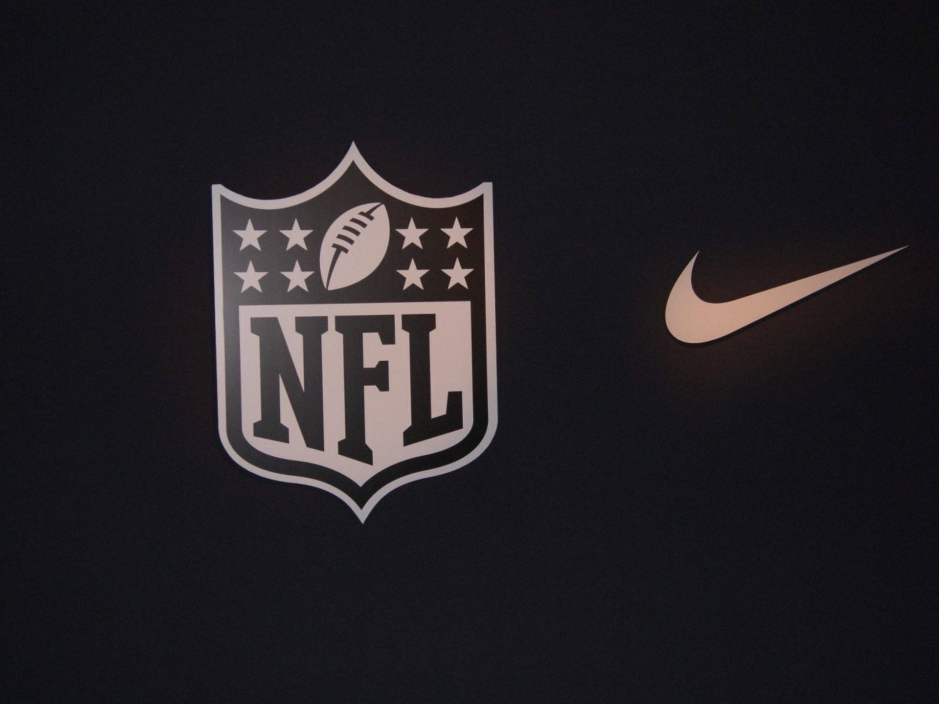 NFL美式足球與Nike品牌標誌 壁紙 — NFL盾牌標誌和Nike勾勾以白色和金色呈現於深色布料背景上。