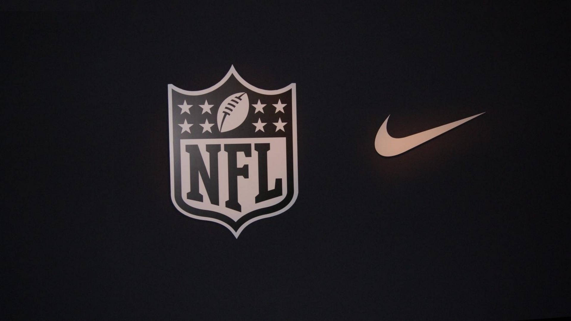 NFL與耐克的經典聯名設計 壁紙 — NFL盾牌標誌和Nike勾勾在深色背景上柔和發光。
