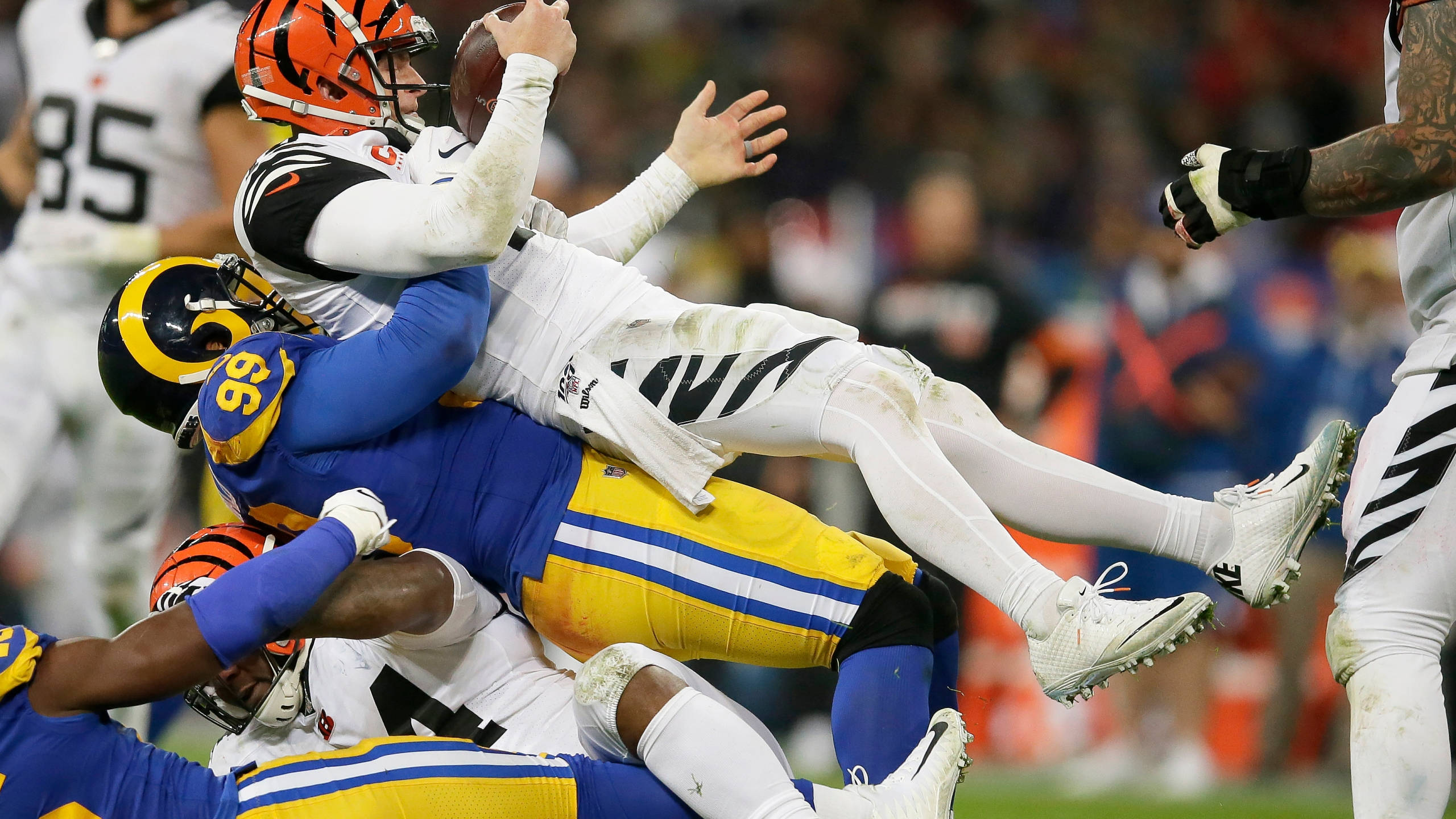 NFL足球員亞倫·唐納德失衡場景 壁紙 — 一名身穿白色球衣的美式足球員在夜間比賽中被藍黃色防守球員擒抱，身體懸空。