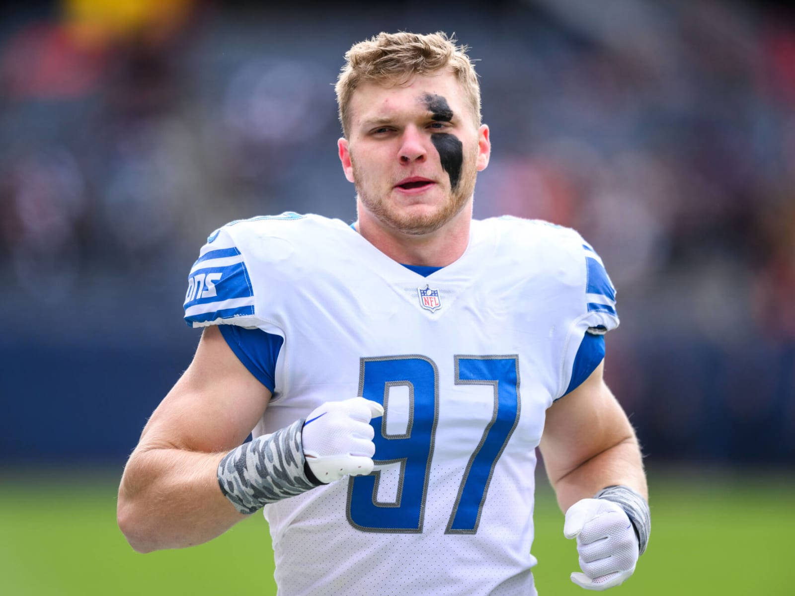NFL防守端鋒艾丹·哈欽遜賽前熱身 壁紙 — 一位決心堅定的NFL球員，身穿白色和藍色球衣，號碼97，帶着黑色眼部塗料，在陽光明媚的場地上衝刺。