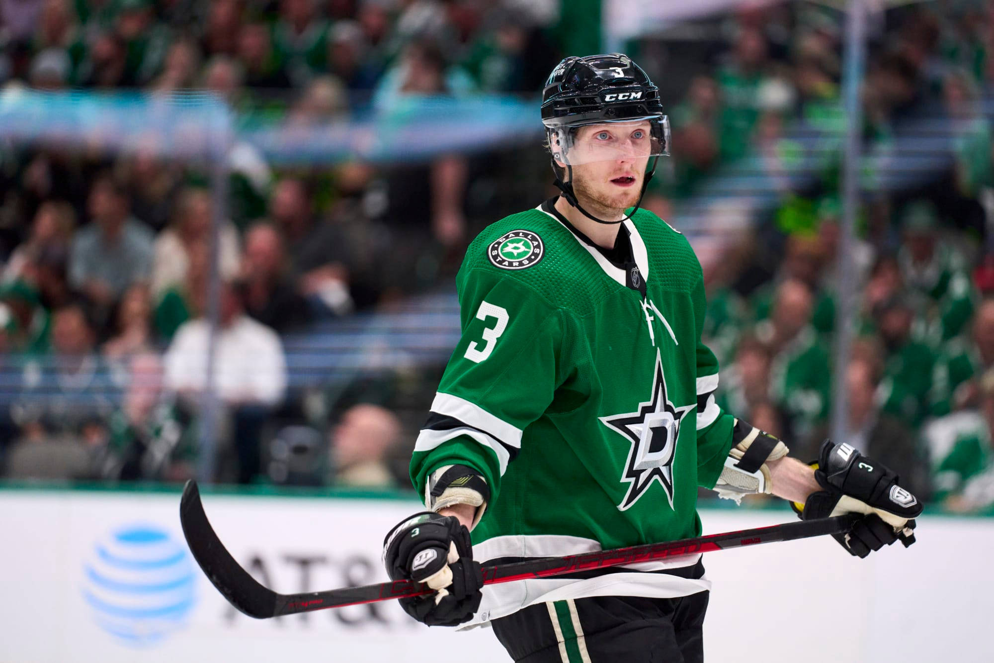約翰·克林格伯格NHL冰球照片 壁紙 — 達拉斯星隊的綠色球衣3號冰球運動員在比賽中站在冰上，手持球桿，背景有觀眾。