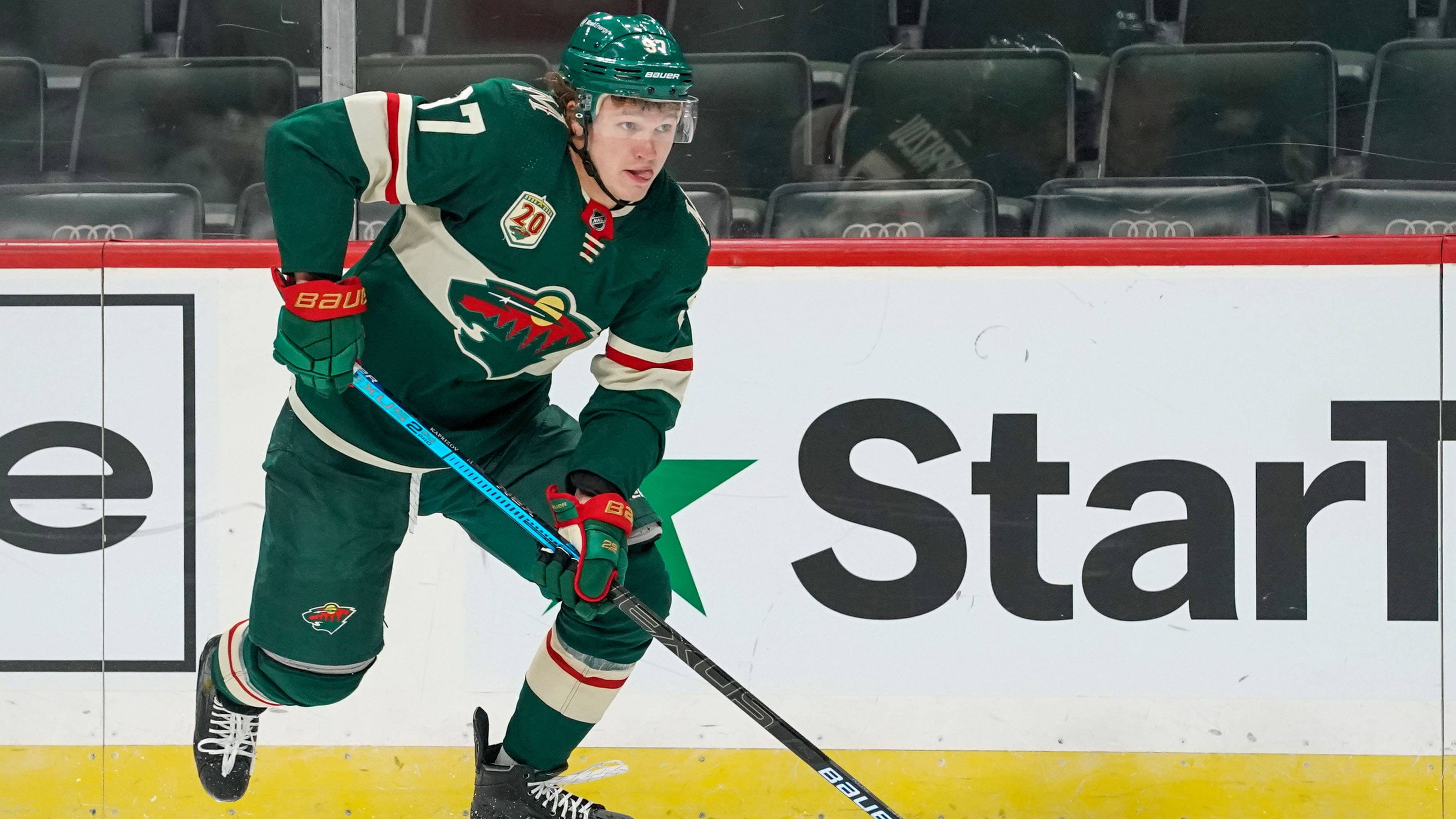 NHL 明尼蘇達狂野隊 基里爾·卡普里佐夫 壁紙 — 一名身穿綠色和白色球衣的冰球運動員在比賽中靠近板牆激烈滑行，背景是空座位。