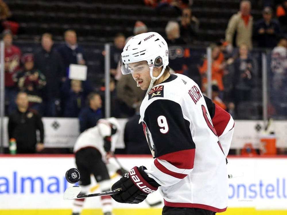 克萊頓·凱勒 NHL比賽 壁紙 — 冰球運動員戴白色頭盔，身穿九號球衣，在冰場上持球棍比賽，背景有模糊的觀眾。