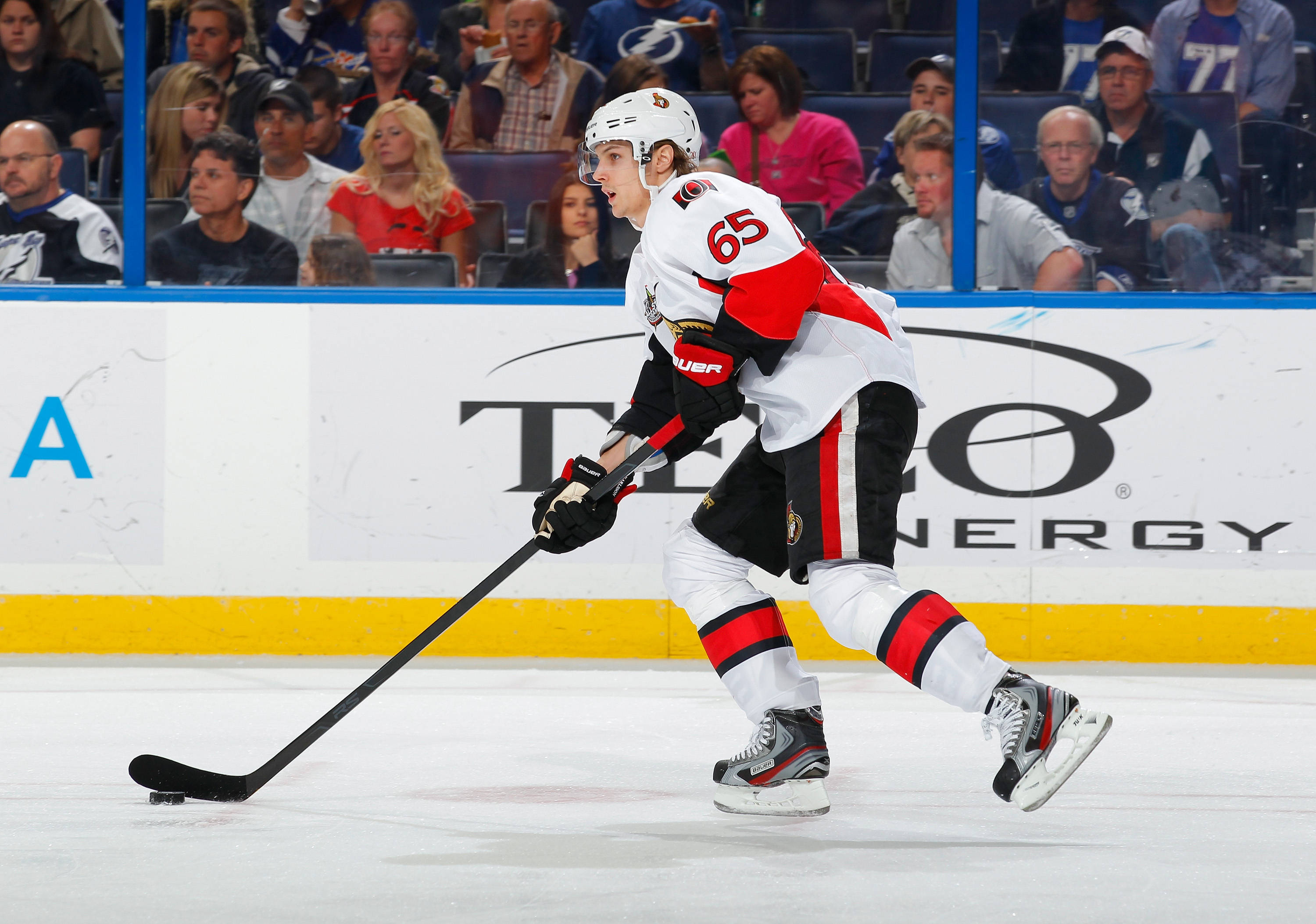 埃里克·卡爾松 NHL 球員冰球渥太華 壁紙 — 一名身穿白色、紅色和黑色球衣的冰球運動員在比賽中手持球棍在冰場上滑行，背景有觀眾。