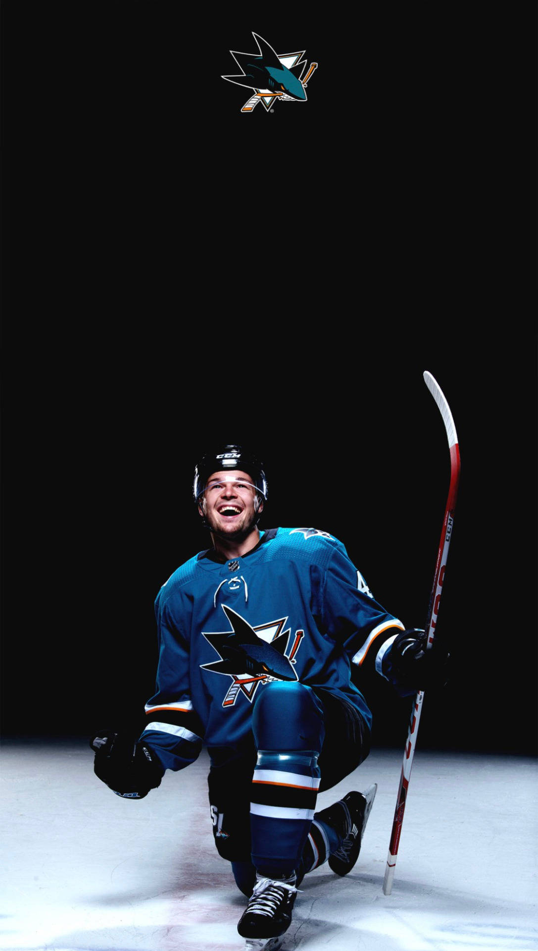 NHL球星托馬斯·赫特爾賽場動態瞬間 壁紙 — 一名身穿聖荷西鯊魚隊球衣的歡欣曲棍球運動員，在黑色背景前跪在冰上，手持球桿。