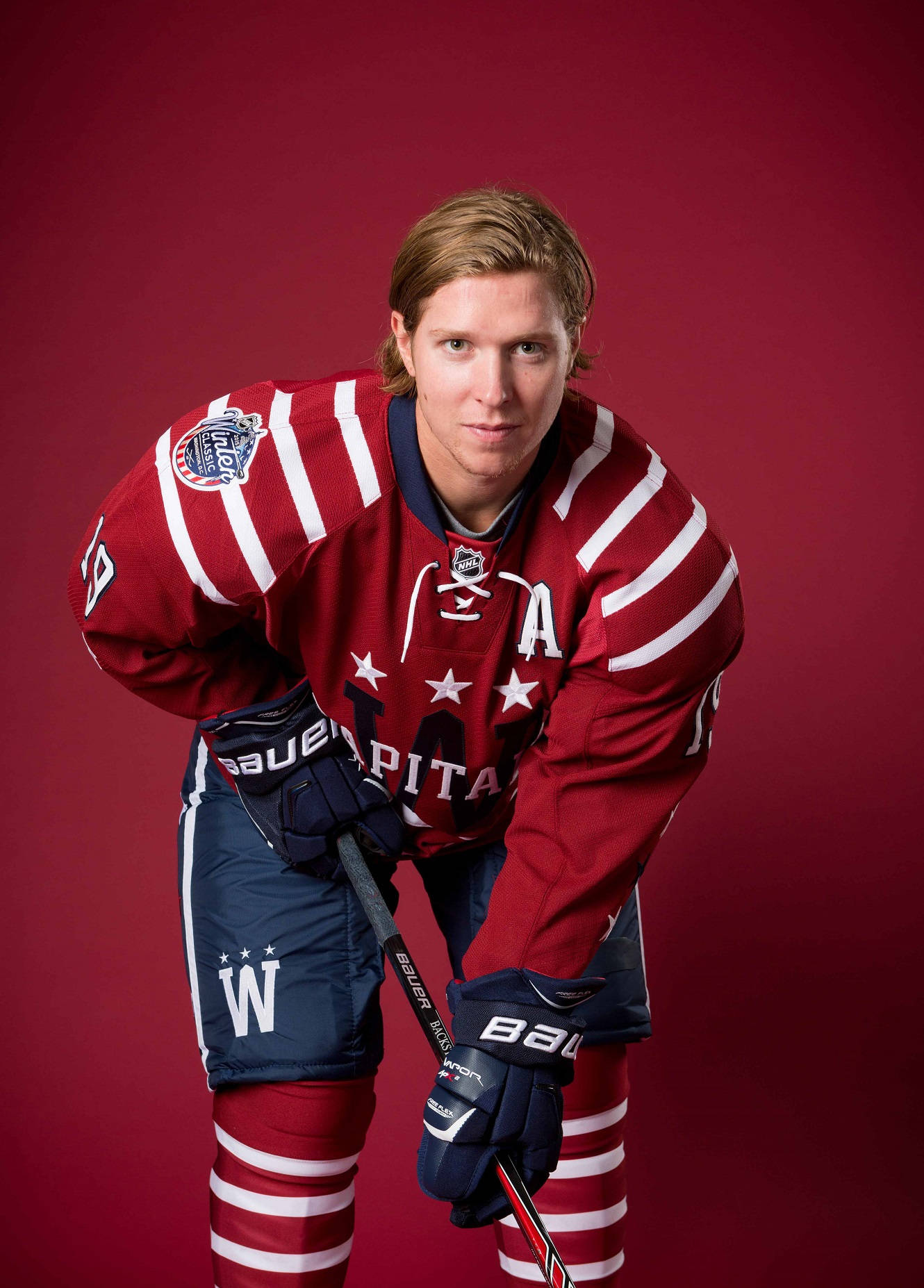 NHL超級巨星尼克拉斯·巴克斯特為華盛頓首都隊出戰 壁紙 — 一名身穿紅白華盛頓首都隊球衣的曲棍球運動員，在純栗色背景前自信地拿著球棒擺姿勢。