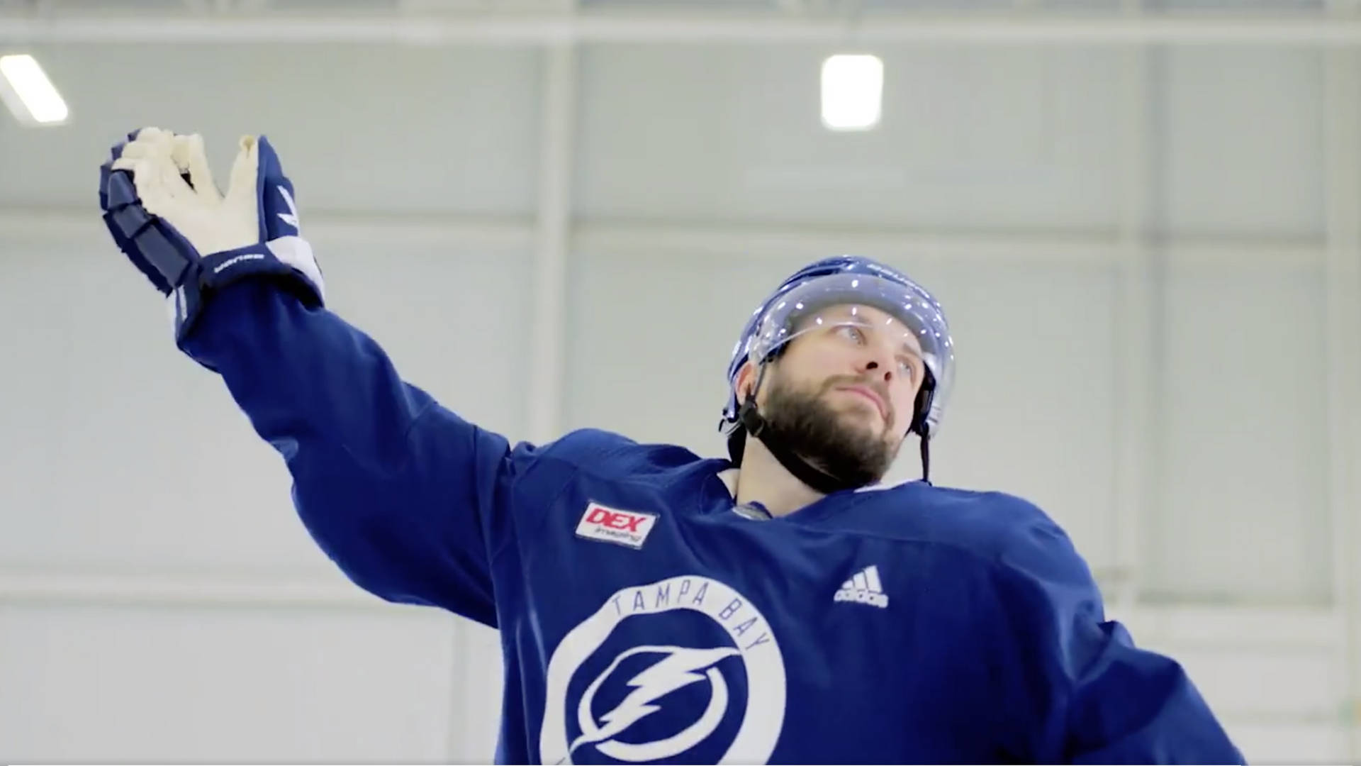 NHL進攻型球員尼基塔·庫察羅夫 壁紙 — 一位身穿藍色裝備的坦帕灣閃電冰球球員，在明亮的燈光下於室內溜冰場舉起戴著手套的手。