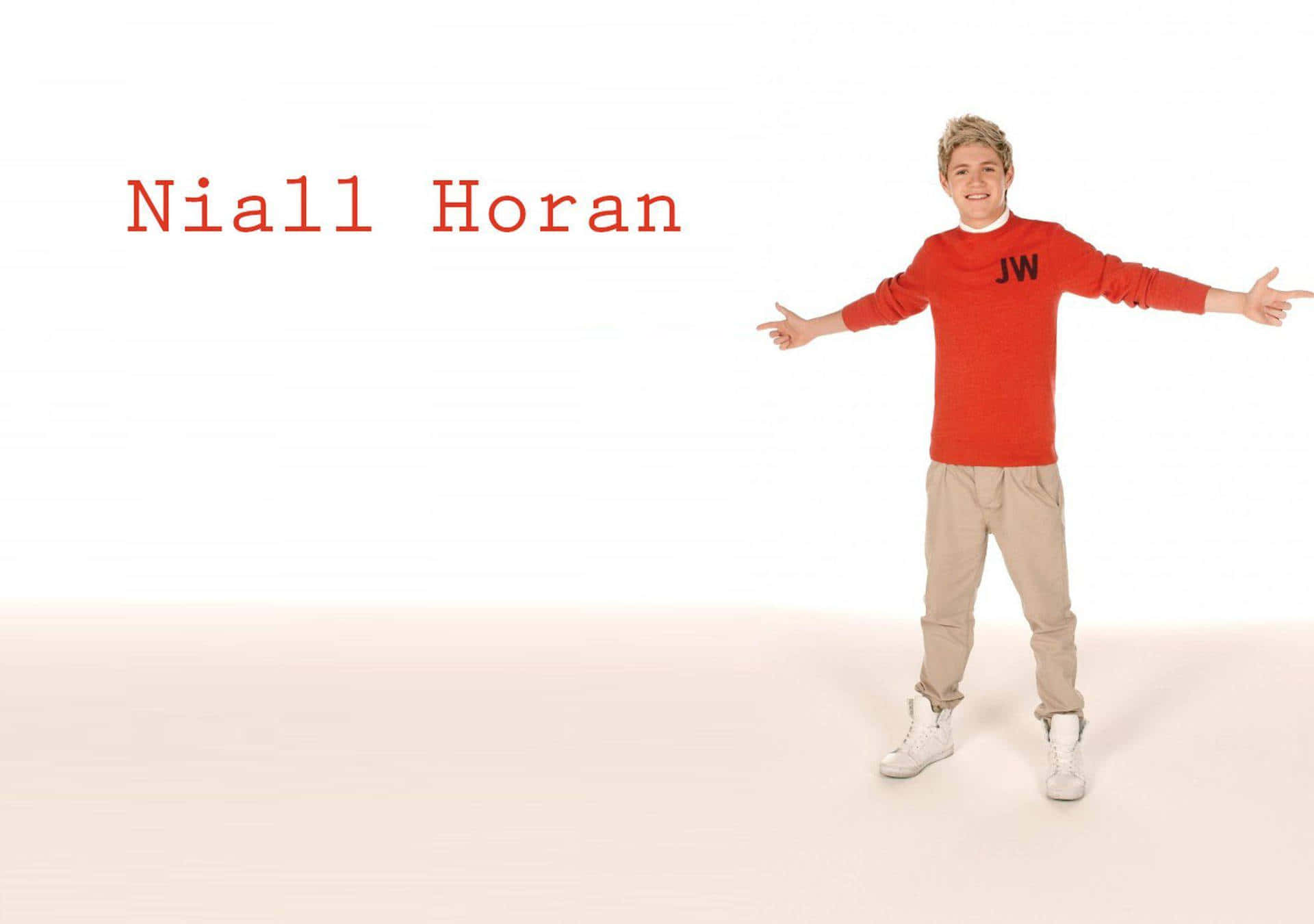  壁紙 — Niall Horan 在白色工作室背景前微笑張開雙臂，身穿紅色毛衣和米色褲子。