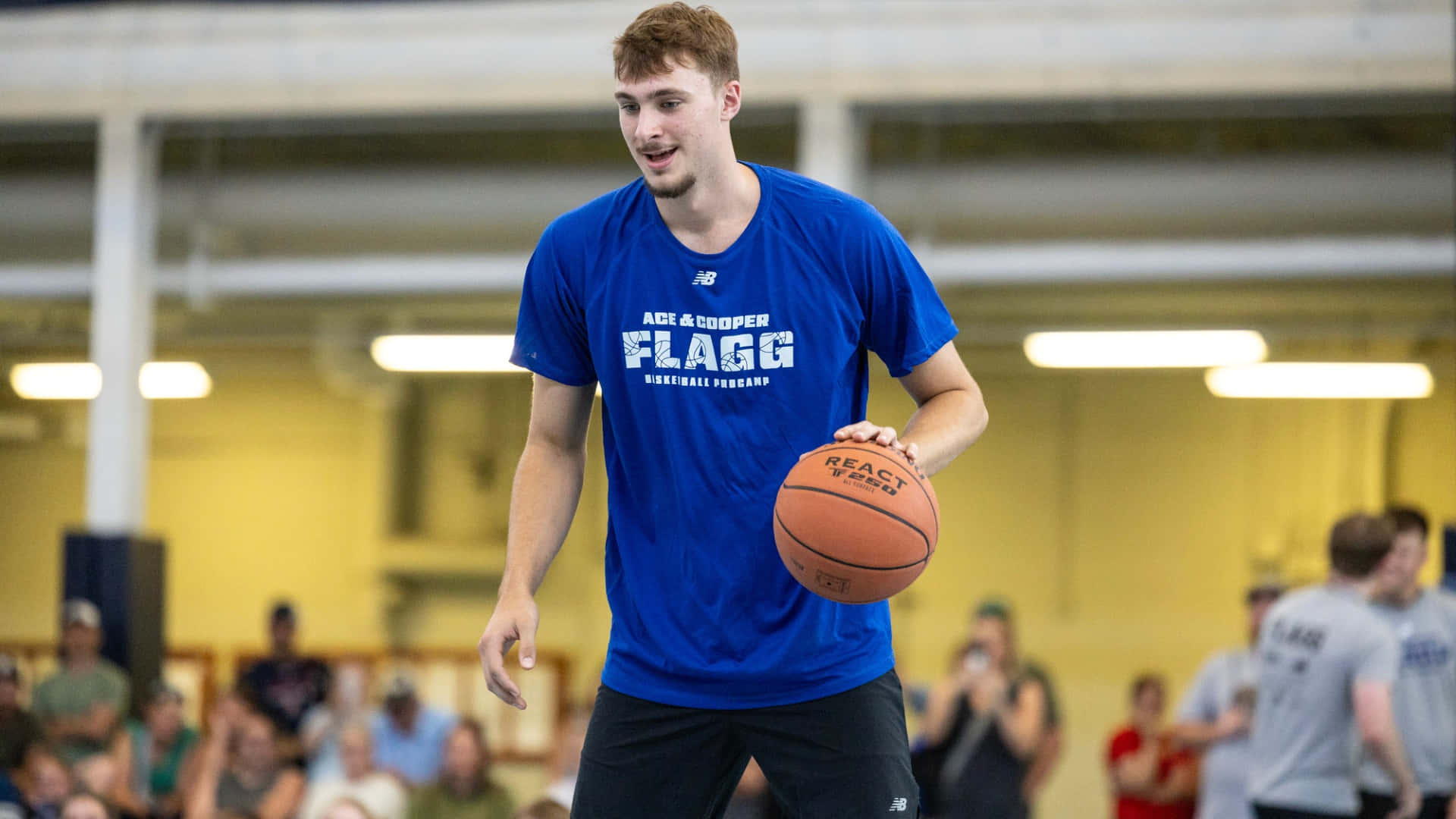  壁紙 — 一名身穿藍色「Flagg Basketball Program」T恤的年輕籃球運動員在室內訓練期間拿著籃球微笑。