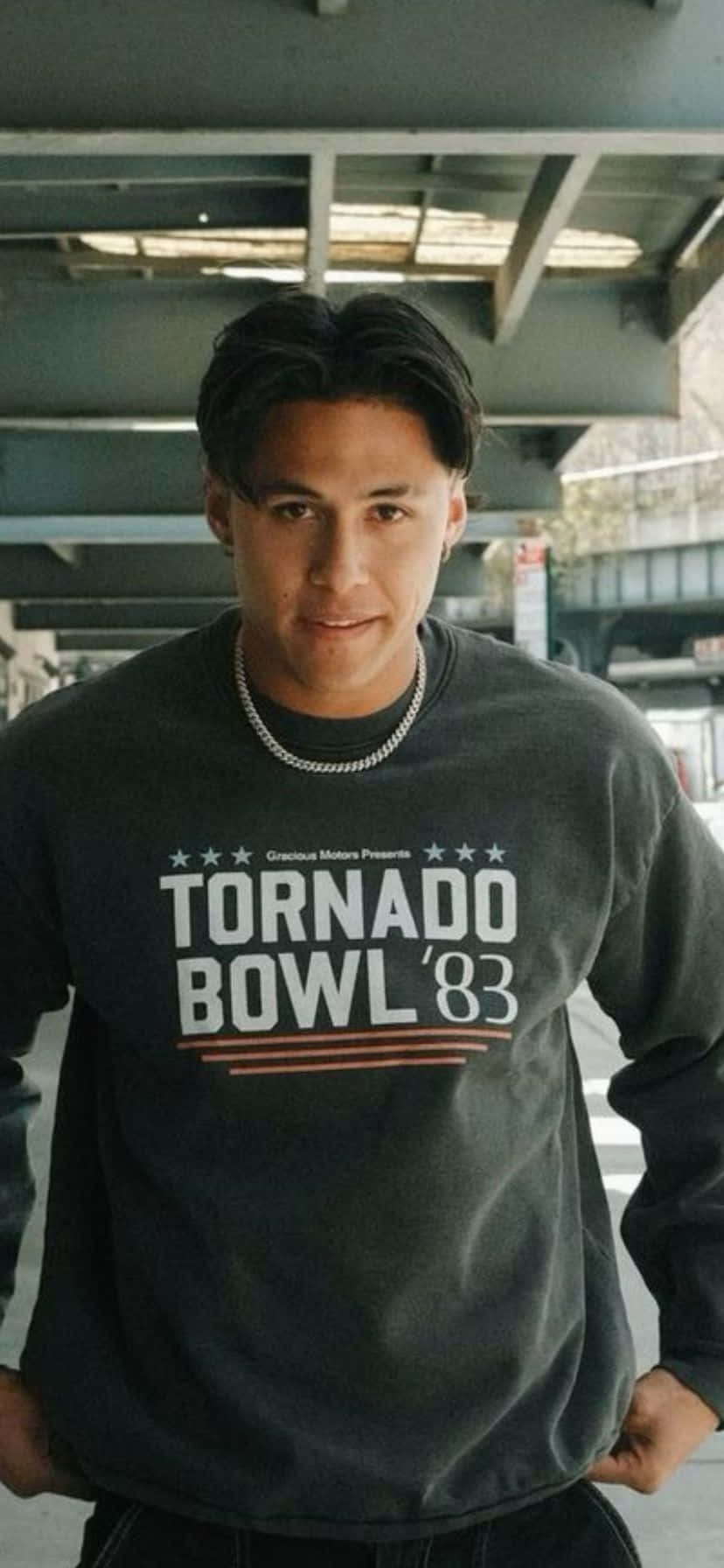 壁紙 — 一名身穿深色衛衣、印有 'TORNADO BOWL '83' 字樣的年輕男子自信地站在城市天橋下。