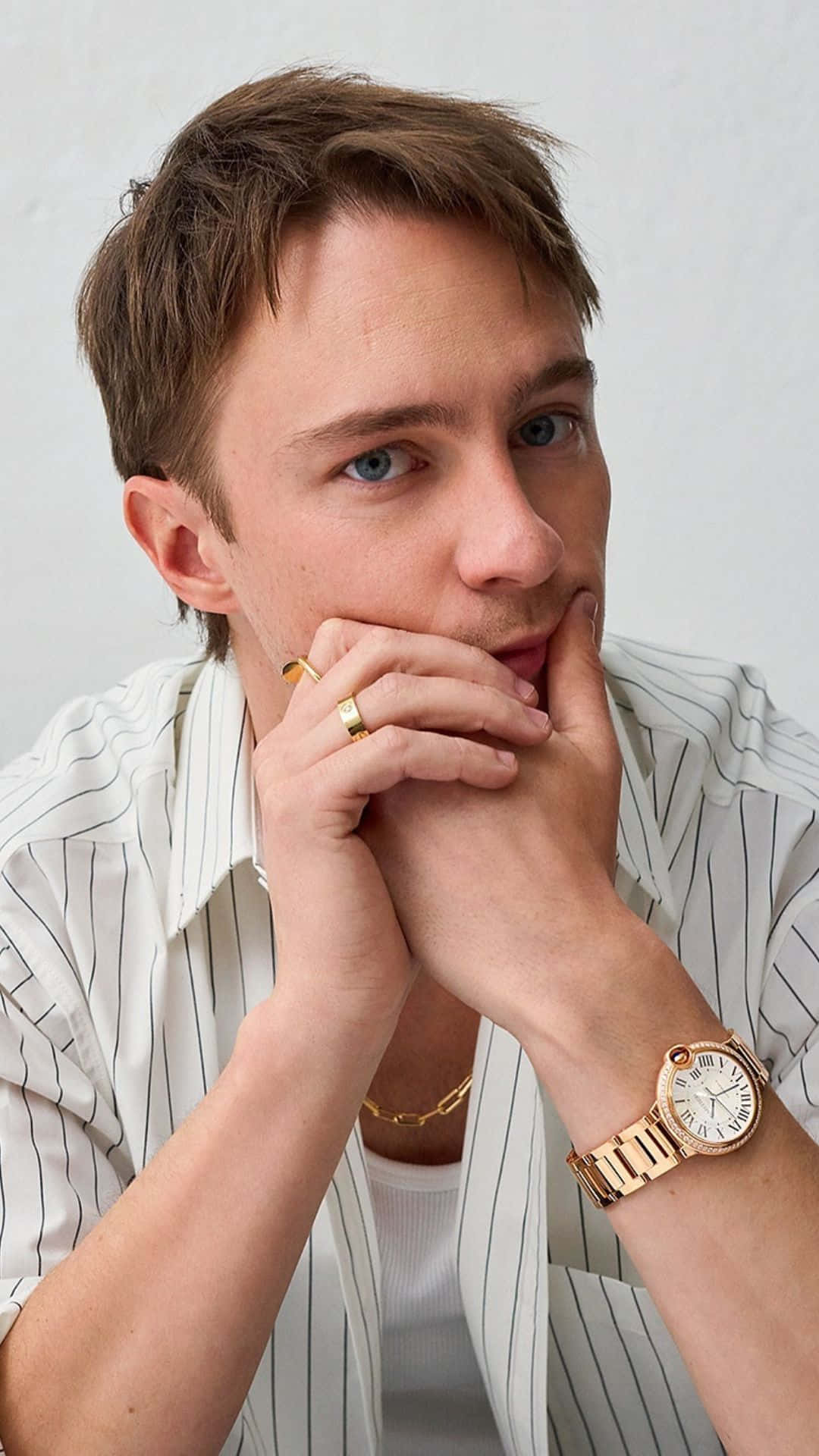  壁紙 — 一位藍眼睛、棕色頭髮的年輕男子穿著條紋襯衫和金色手錶，雙手交疊托著下巴，靠在一面純白牆上。