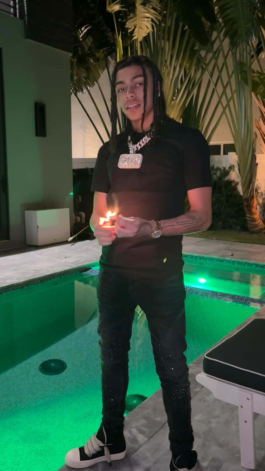 壁紙 — 一位留著辮子的年輕男子夜晚站在發光的綠色泳池旁，手持點燃的蠟燭，穿著黑色閃亮褲子和鏈條項鍊。
