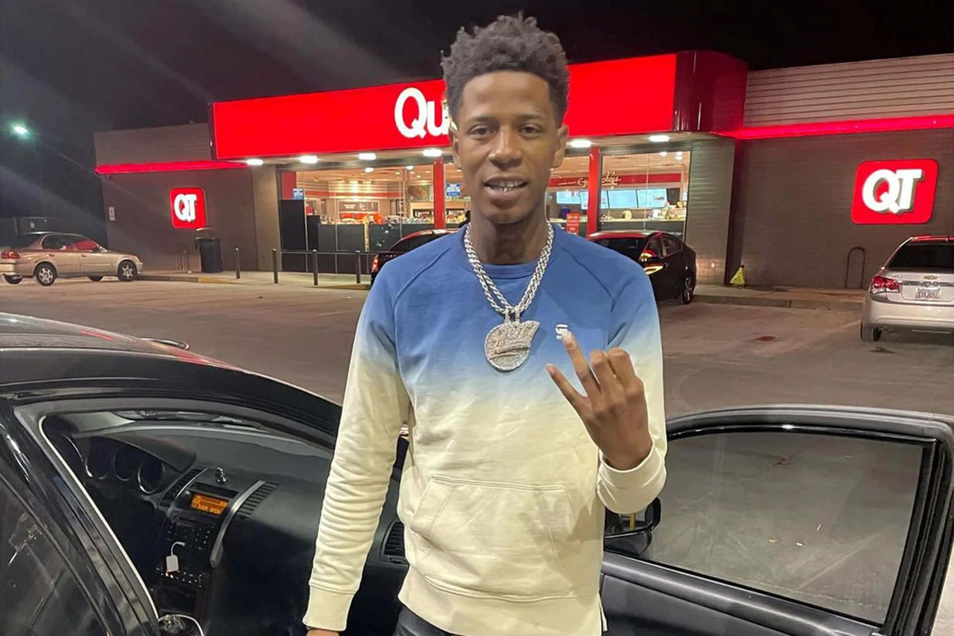  壁紙 — 一名身穿藍色和奶油色運動衫的年輕男子，在夜間自信地站在一輛汽車旁，身後是燈火通明的QT加油站。