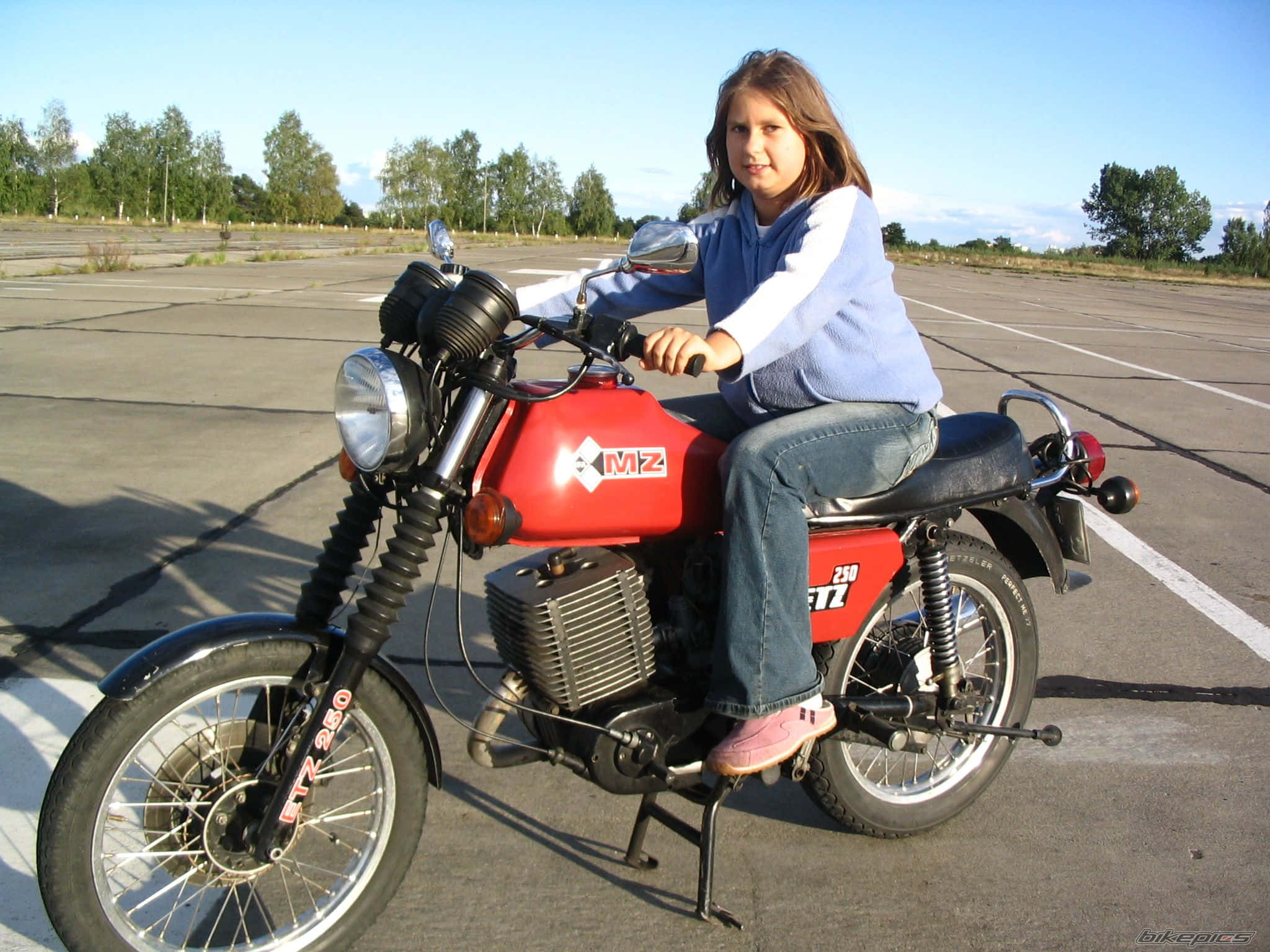 Mz Motorcycle 桌布 壁紙 — 一個年輕女孩自信地坐在一輛紅色復古電單車上，背景是陽光普照的停車場和樹木。