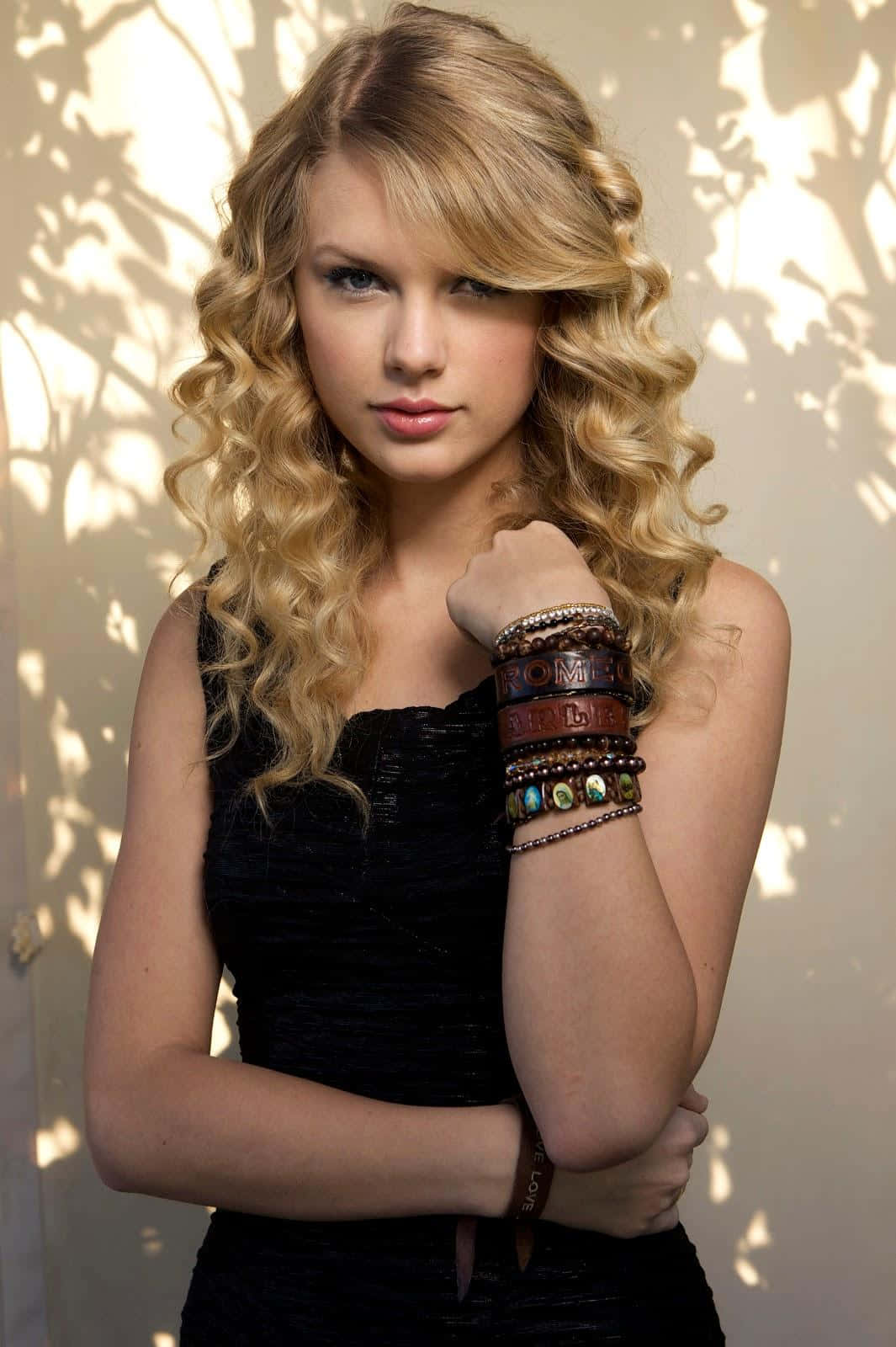 Taylor Swift Lover 桌布 壁紙 — 一位年輕女性擁有豐盈的金色捲髮，穿著黑色上衣和疊戴的皮革手鐲，背景是陽光照射的牆壁和樹葉陰影。