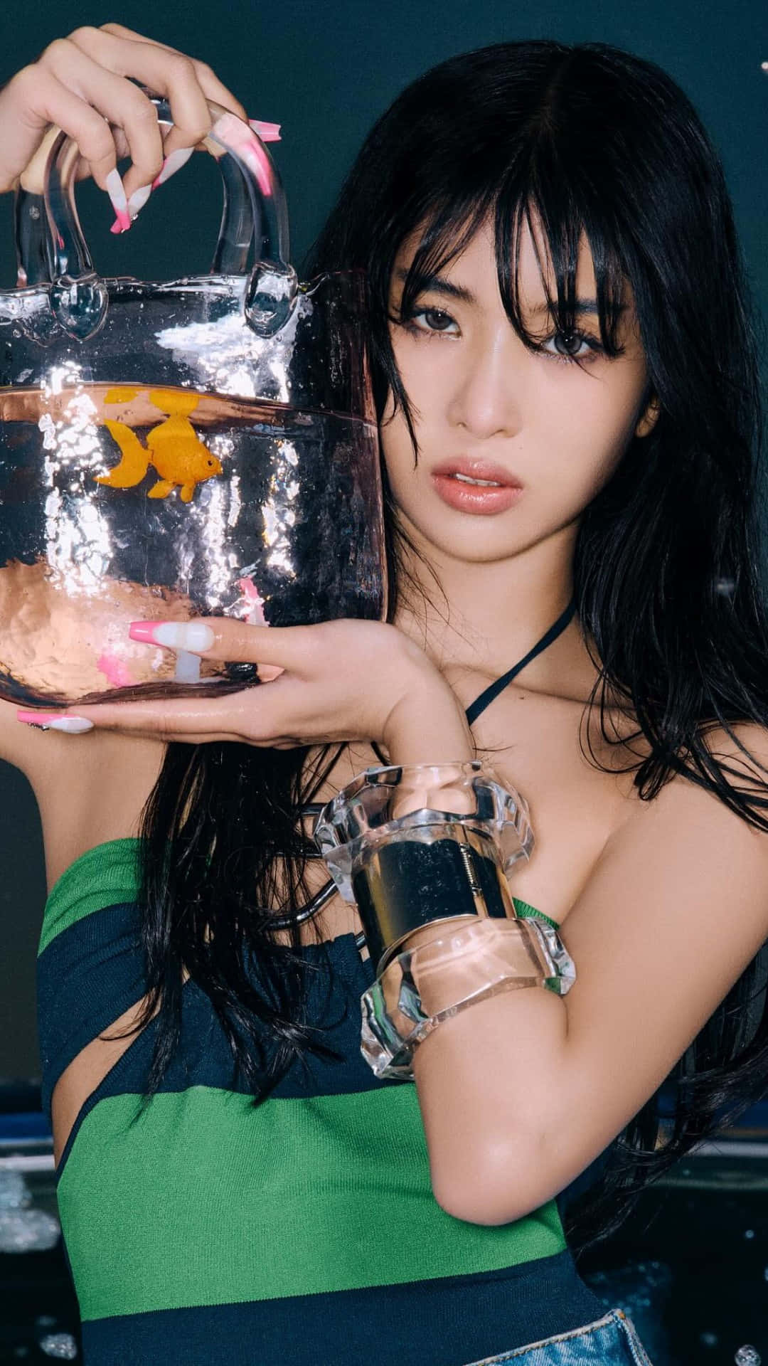  壁紙 — 一位深色頭髮的年輕女性手持一個透明手袋，內有金魚和水，佩戴大膽的配飾和綠色上衣。