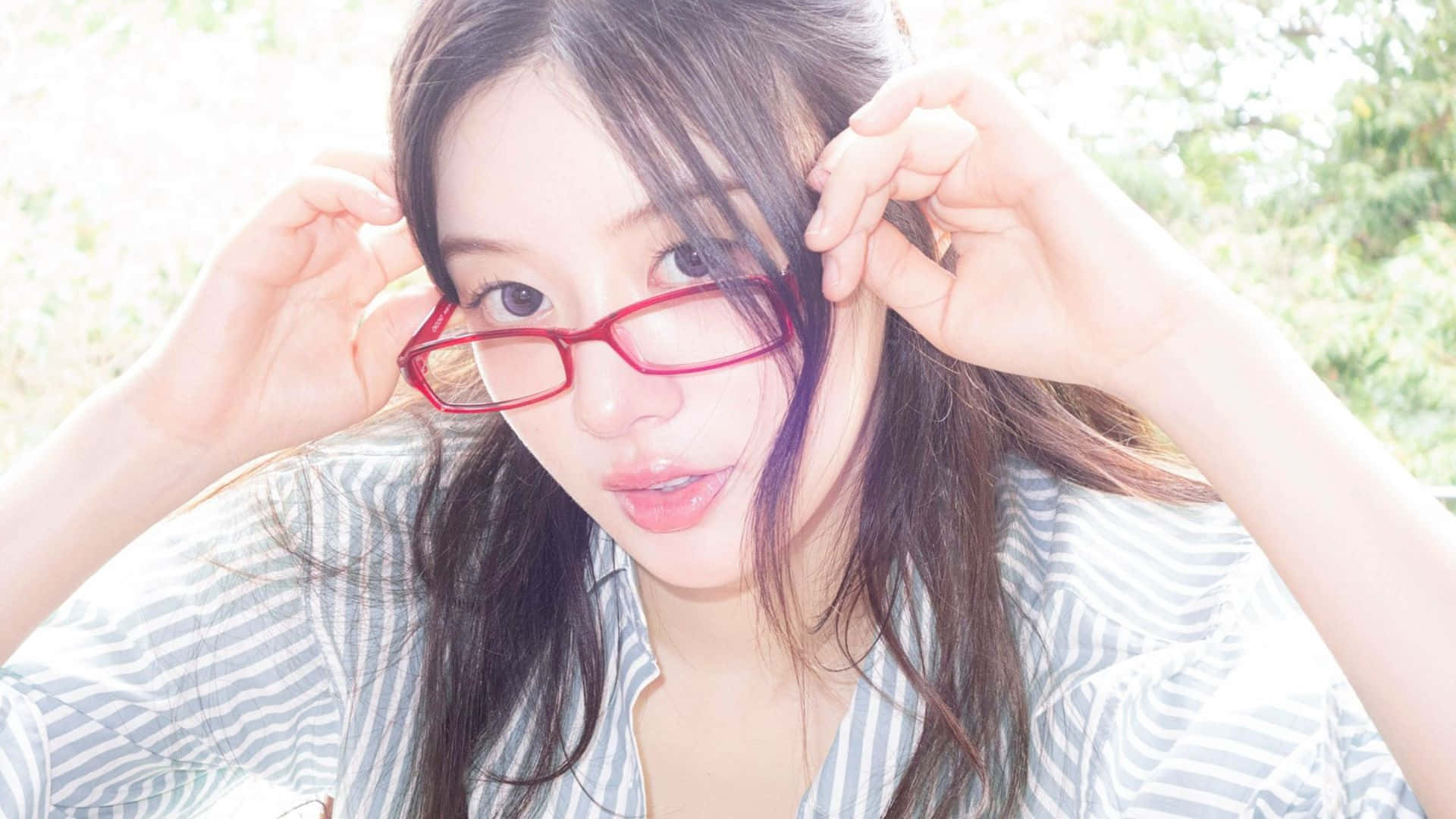  壁紙 — 一位深色頭髮的年輕女性在柔和日光下調皮地舉起紅色框眼鏡，同時直視鏡頭。