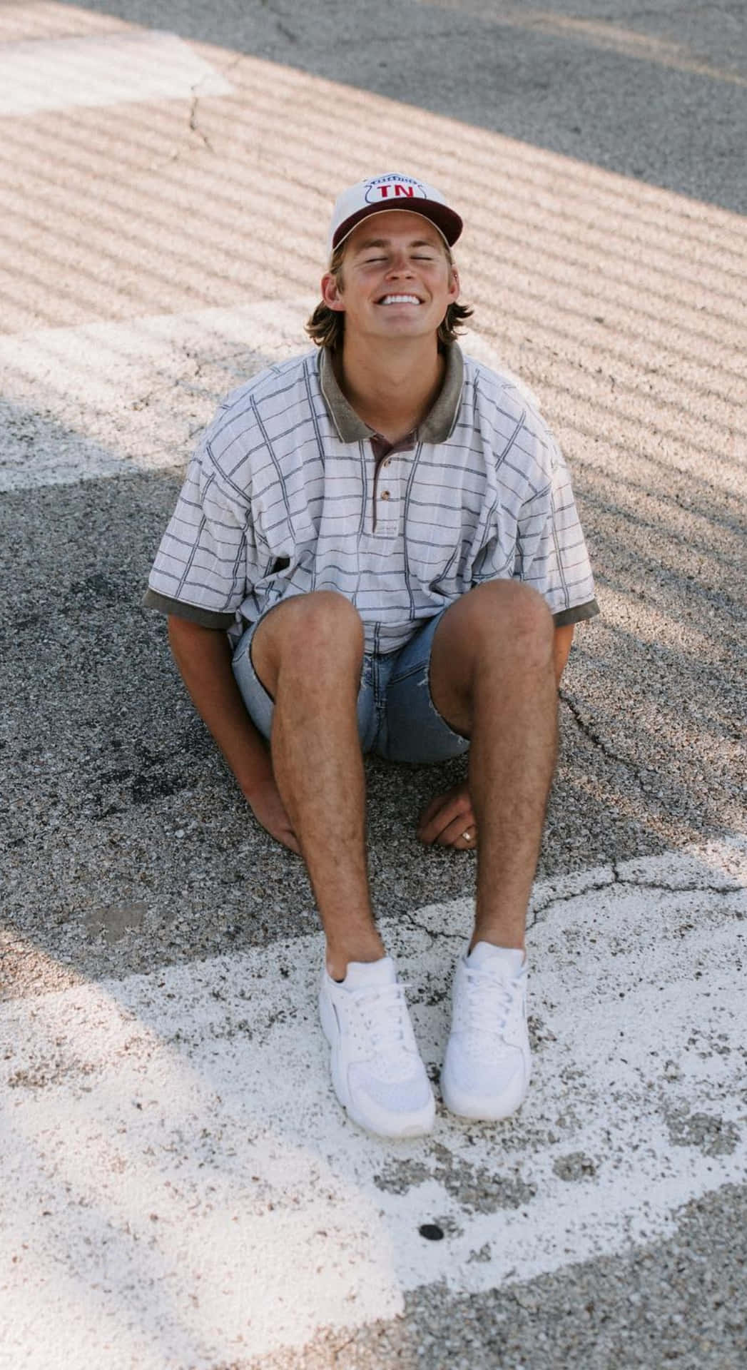  壁紙 — 一個年輕男子帶著愉快的笑容，盤腿坐在陽光普照的瀝青路上，戴著田納西帽和白色運動鞋。