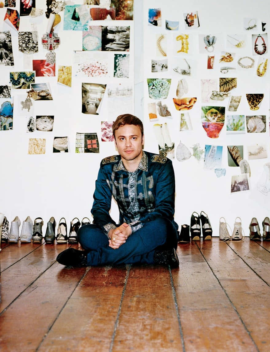 Nicholas Kirkwood為《Vogue》品牌意識專頁拍攝 壁紙 — 一個男人盤腿坐在木地板上，周圍是鞋子，牆上覆蓋著不拘一格的拼貼藝術。