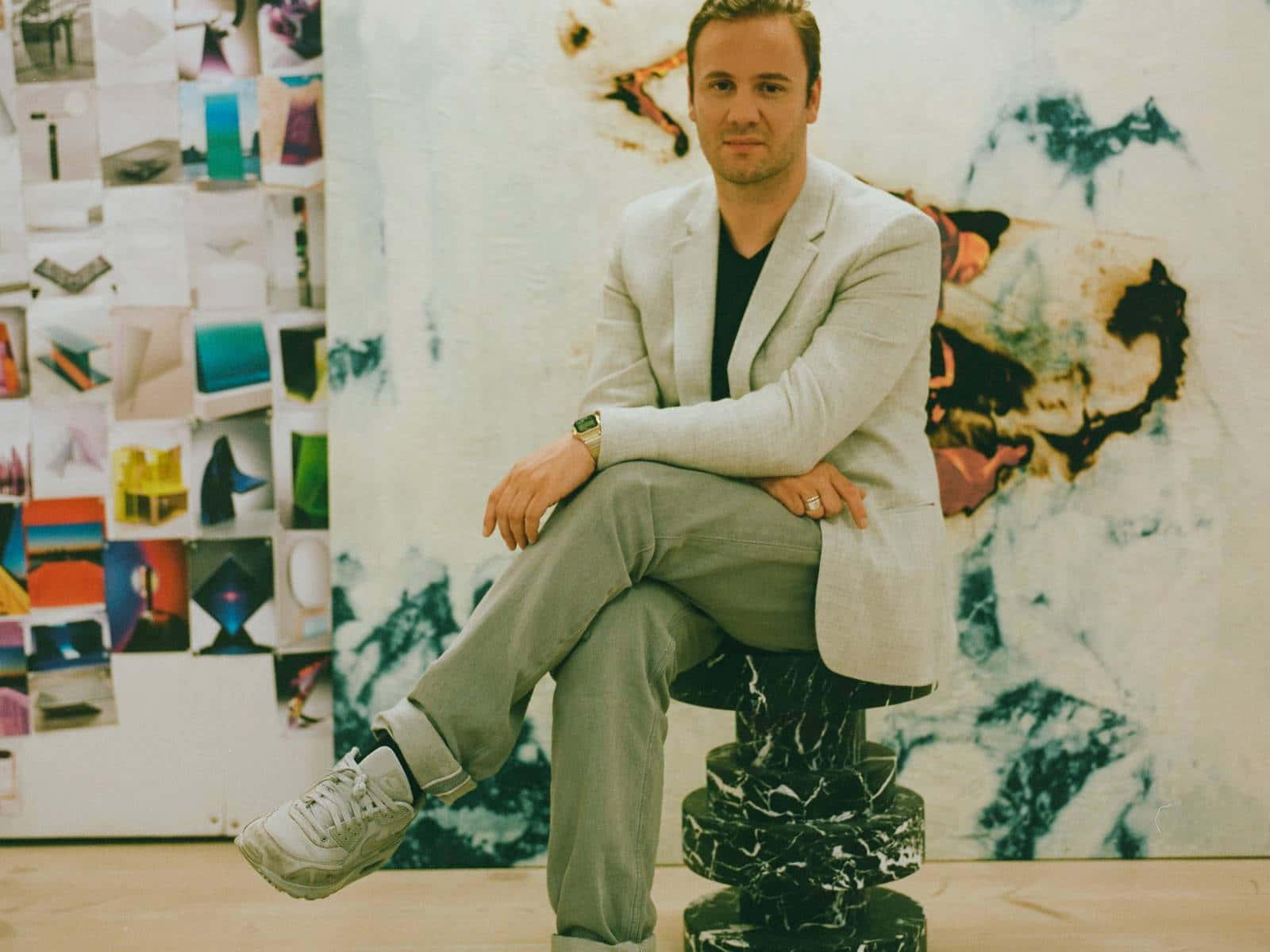 Nicholas Kirkwood倚靠抽象牆 壁紙 — 一名身穿淺色西裝外套的男子，隨意地坐在黑色大理石凳上，背景是抽象藝術和拼貼牆。