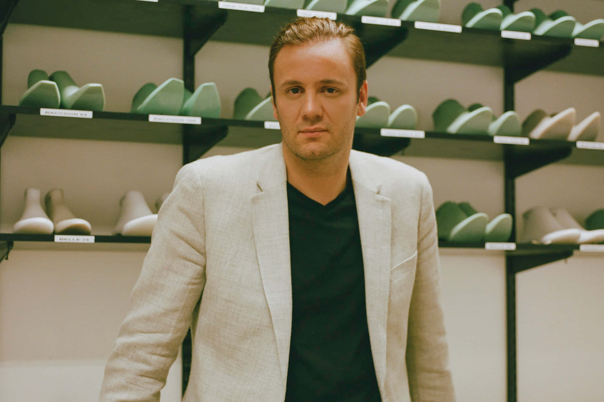 Nicholas Kirkwood站在鞋櫃前 壁紙 — 一位身穿淺色西裝外套的男子自信地站在鞋店裡，身後是擺滿綠色和白色鞋子的貨架。