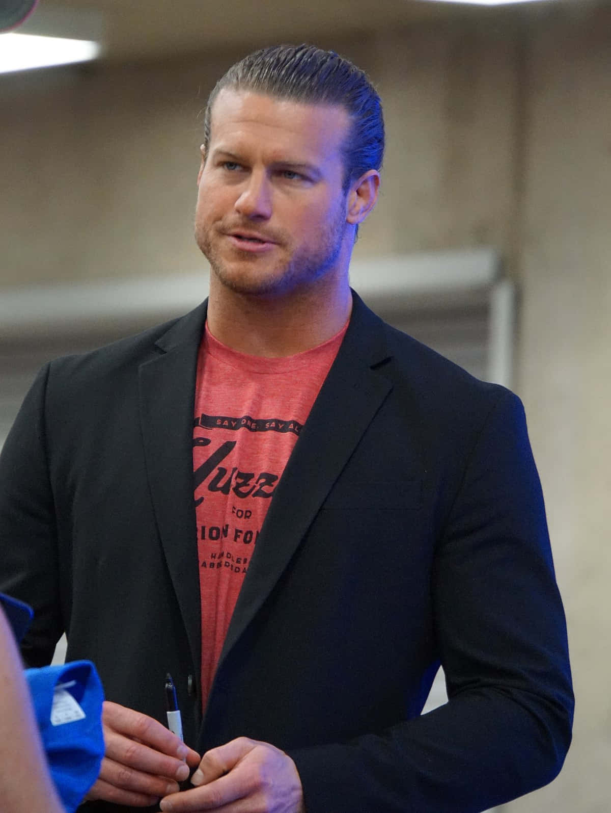 Nicholas Theodore Nemeth（即Dolph Ziggler） 壁紙 — 一個肌肉發達、頭髮向後梳的男人，身穿黑色西裝外套內搭紅色T恤，在室內拿著一支筆。