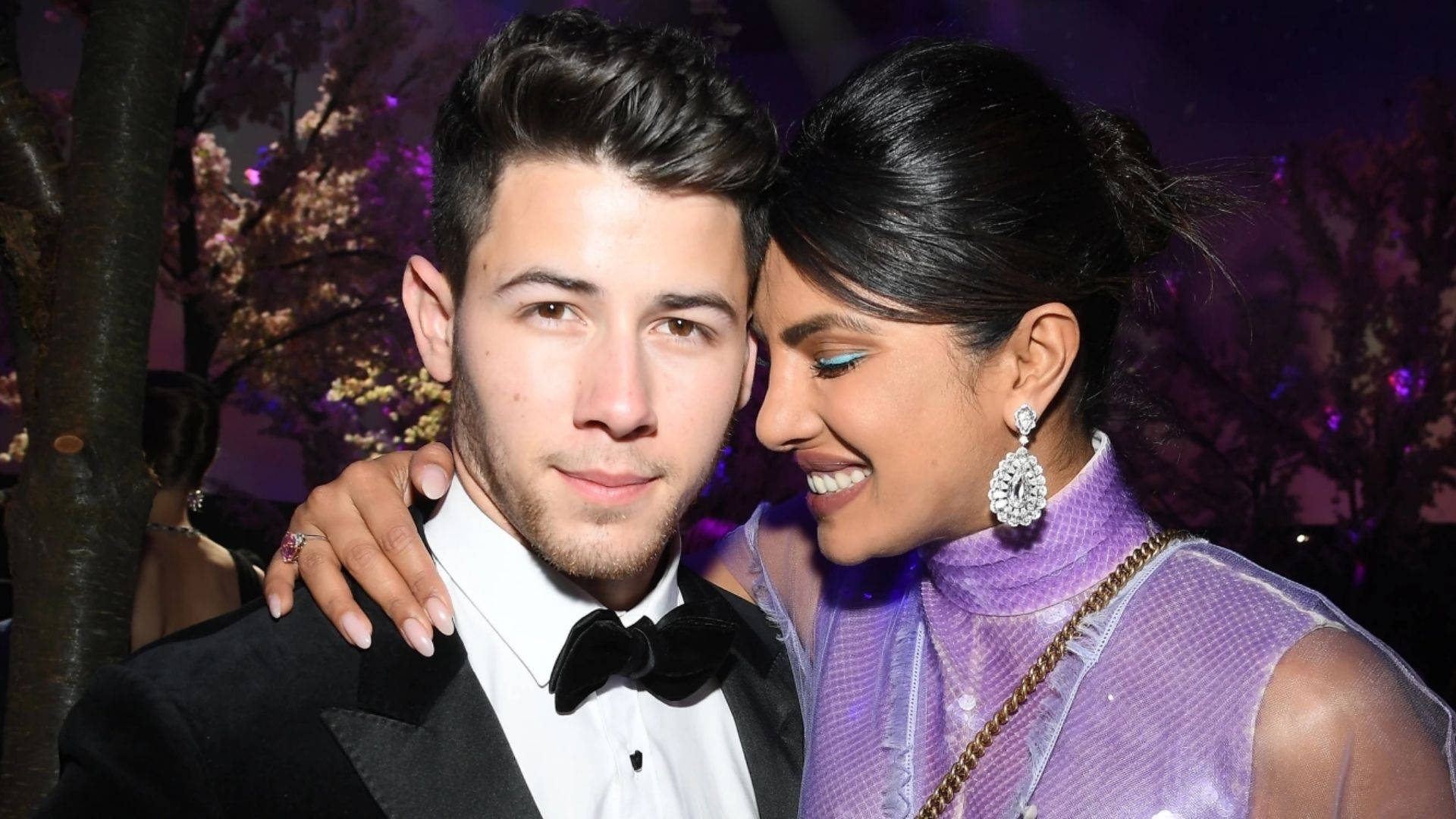 Nick Jonas 與妻子 壁紙 — 一對微笑的情侶在晚間活動中緊密地擺姿勢，背景有紫色燈光和盛開的樹木。