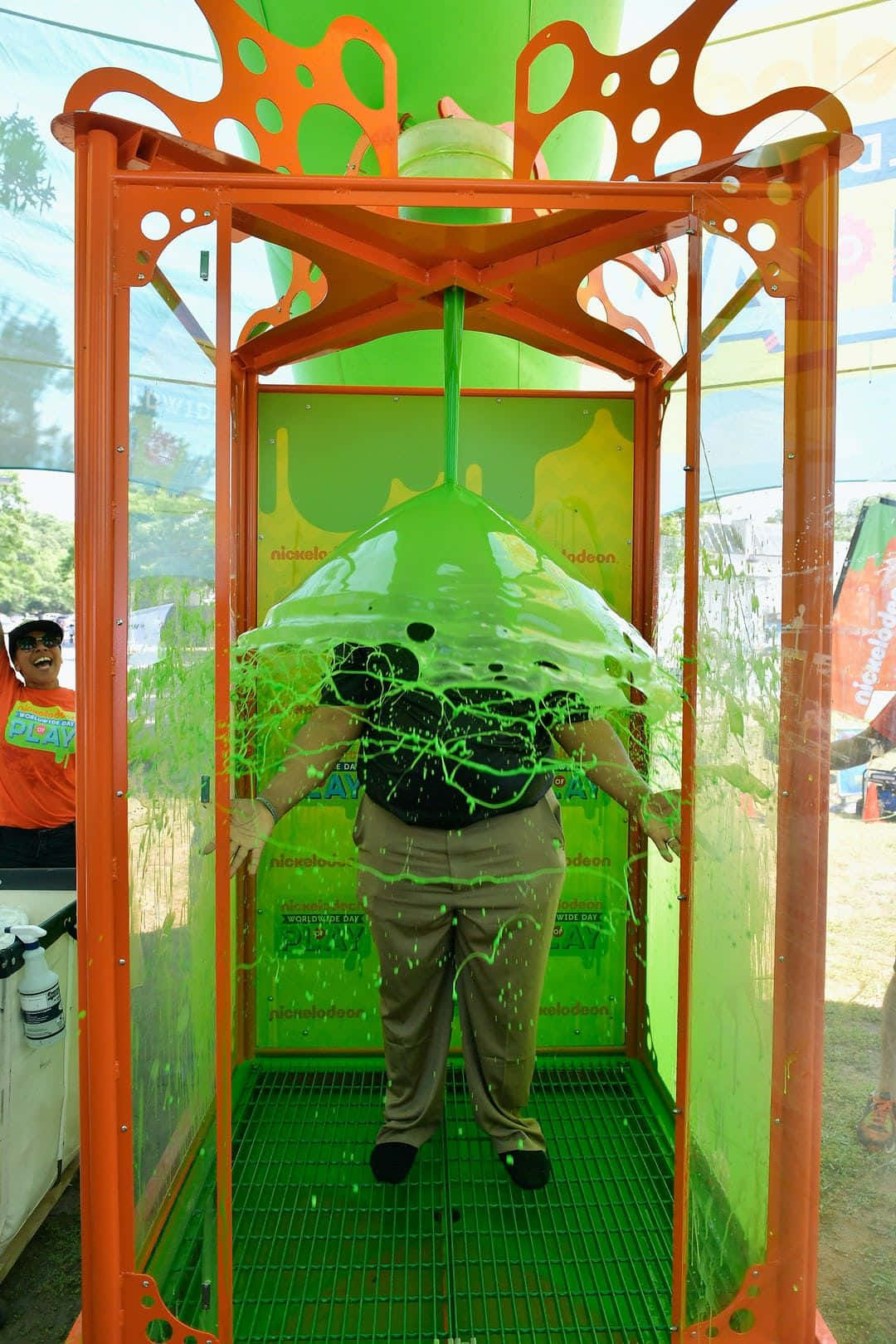 Nickelodeon史萊姆圖片 壁紙 — 一個人站在一個鮮橙色嘅黏液大炮攤位入面，綠色黏液喺戶外活動中濺到佢哋身上。