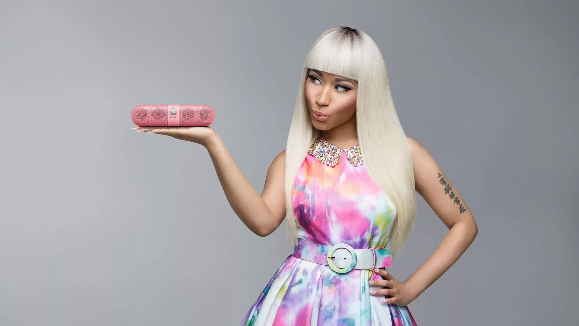 Nicki Minaj 白髮造型 壁紙 — 一位擁有鉑金色頭髮、身穿彩色連衣裙的女性，在純灰色背景前拿著一個粉紅色便攜式揚聲器。