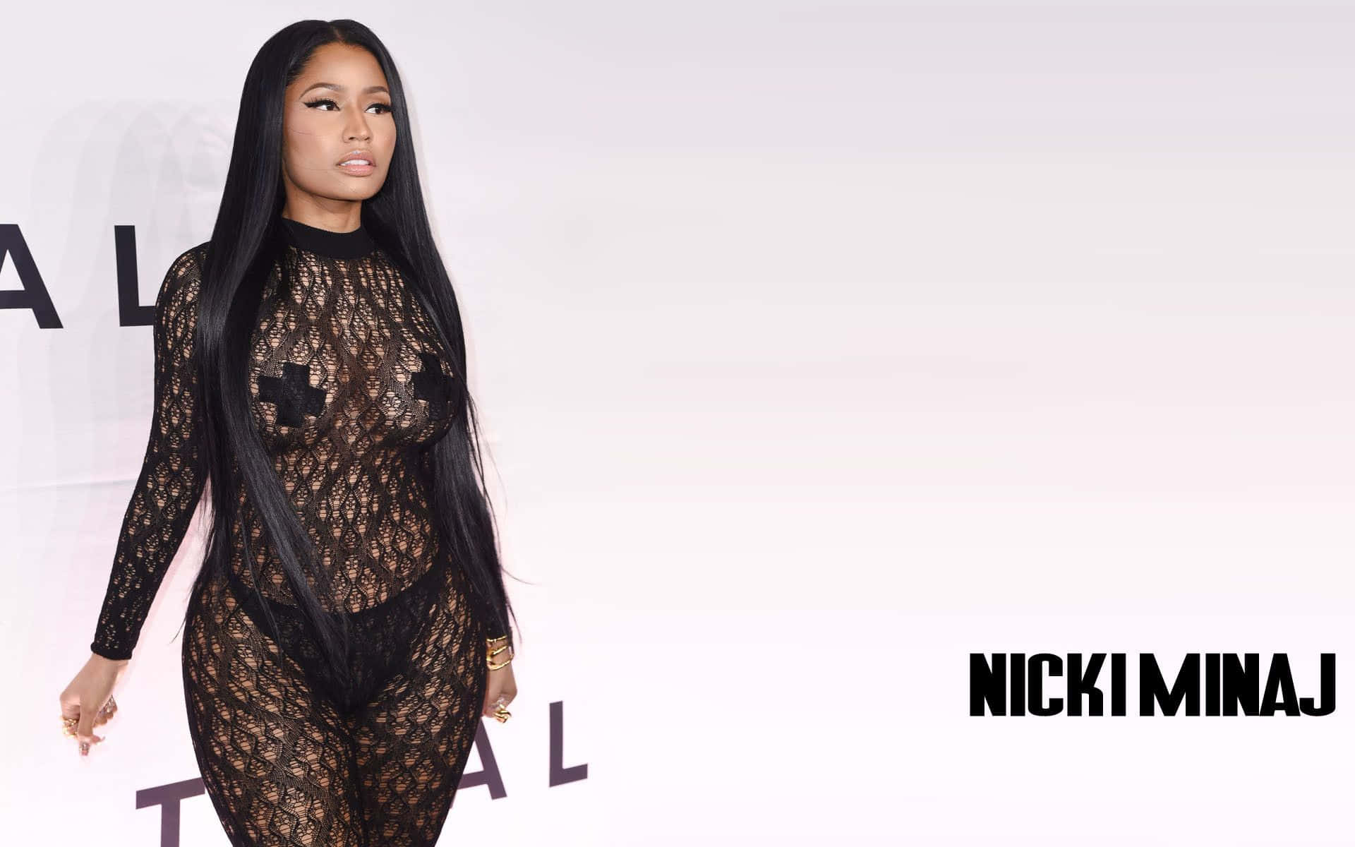 妮琪·米娜 桌布 壁紙 — Nicki Minaj 自信地穿著透視黑色蕾絲連體衣，在極簡白色背景前行走，並顯示她的名字。